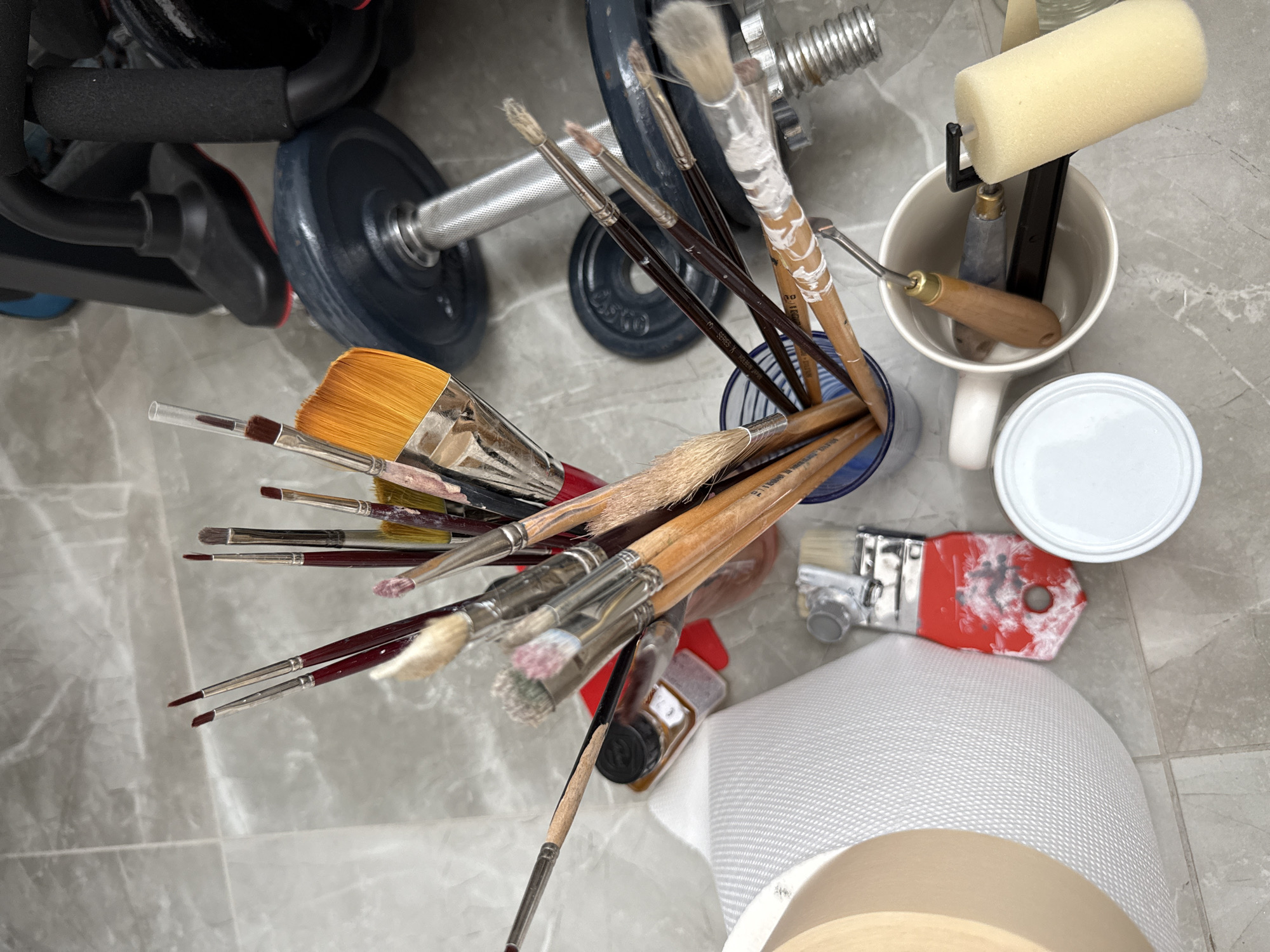 The image depicts an overhead view of art supplies and workout equipment arranged on a tiled floor. In the foreground, a collection of paintbrushes in various sizes and states of use are held in a blue cylindrical container. Some brushes are clean, while others show traces of paint. Adjacent to the container, a white roll of textured material, possibly canvas or paper, is partially visible on the left. To the right of the brushes, a white mug contains several sculpting tools and a small foam roller. A white lid for a container is also present near the mug. In the background, weightlifting equipment, including dumbbells and part of a weight machine, are visible. A small bottle with a black cap and amber liquid, possibly paint or varnish, is positioned among the paintbrushes. The scene appears to be set in a home studio or workshop environment in Mesogi, Cyprus. There are no people or active interactions visible in the image. The lighting suggests an indoor setting, possibly with natural light from above. The floor is made of grey tiles with subtle veining.