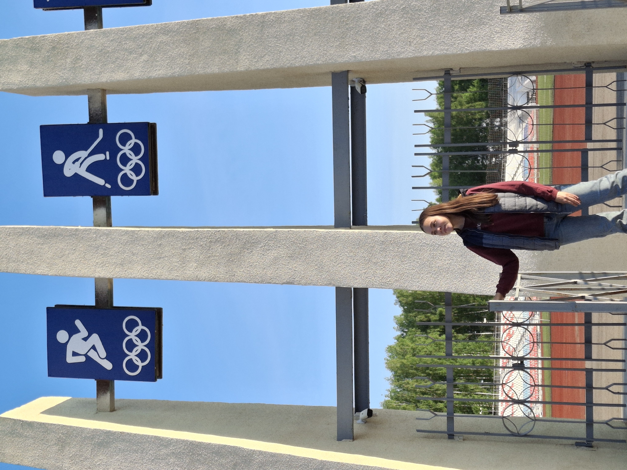 A girl with long brown hair is looking over a fence. Behind the fence is a stadium, which is partially visible through the fence. In the background are trees and part of the stands. To the left of the fence are two signs with a white figure falling down, alongside the Olympic rings, which indicate the location as being the Chaikovsky Olympic Training Center in Russia. The sky is blue. The picture is taken at an odd angle.