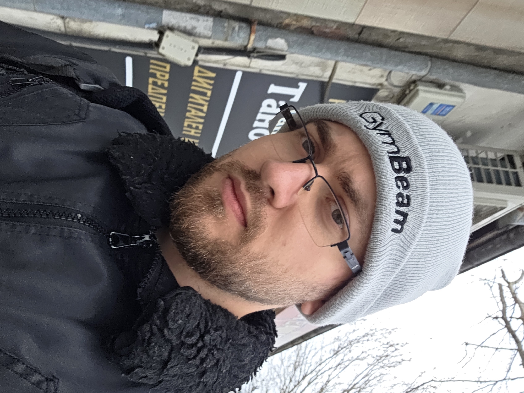 A man wearing a gray "GymBeam" beanie and glasses poses for a selfie outdoors. He is wearing a black jacket with a fur collar. Behind him, there is a sign with Cyrillic script, likely indicating a business in Plovdiv, Bulgaria. Utility boxes and bare trees suggest an urban setting, possibly a street scene in winter.