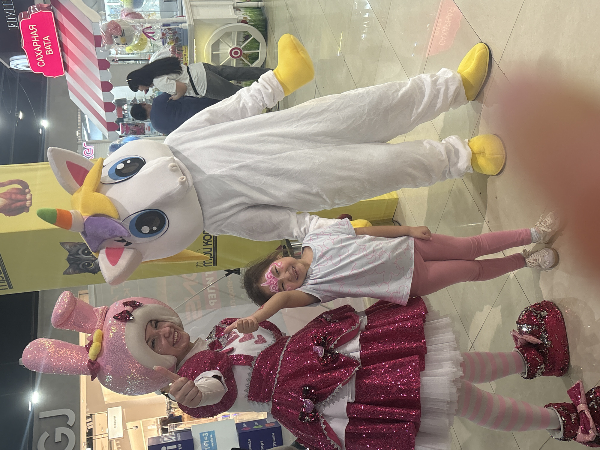 Here's a detailed description of the image:
**Setting:** An indoor commercial space, likely a shopping mall or retail area in Tri Potoka, Russia, judging by the Cyrillic writing visible.
**People:** The image features:
* Two individuals in full-body character costumes: one resembling a white unicorn and another resembling a pink bunny.
* A young girl with face paint.
* At least two adults are visible in the background.
* The costumed individuals are interacting with the young girl, likely entertaining her.
**Subjects & Objects:**
* **Costumes:** The unicorn costume is white with yellow accents (mane, shoes). The bunny costume is pink with silver glitter details, a white ruffle, and pink and white striped stockings.
* **Child:** The girl has a face paint design on her cheek. She wears a light-colored patterned t-shirt and pink leggings.
* **Background elements:** Visible are store displays, including a sign with Cyrillic text ("САХАРНАЯ ВАТА"), shelving with toys and clothing.
* Signs include Cyrillic text: "ДЕВУШКИ", "1+1=3 на обувь".
**Actions & Interactions:**
* The child is interacting directly with both costumed performers. She is smiling and engaging with them.
* The costumed performers appear to be posing for a picture.
**Environment:**
* The flooring is a light-colored tile.
* The lighting is bright and artificial.
* The scene gives the impression of a promotional event within a retail environment.