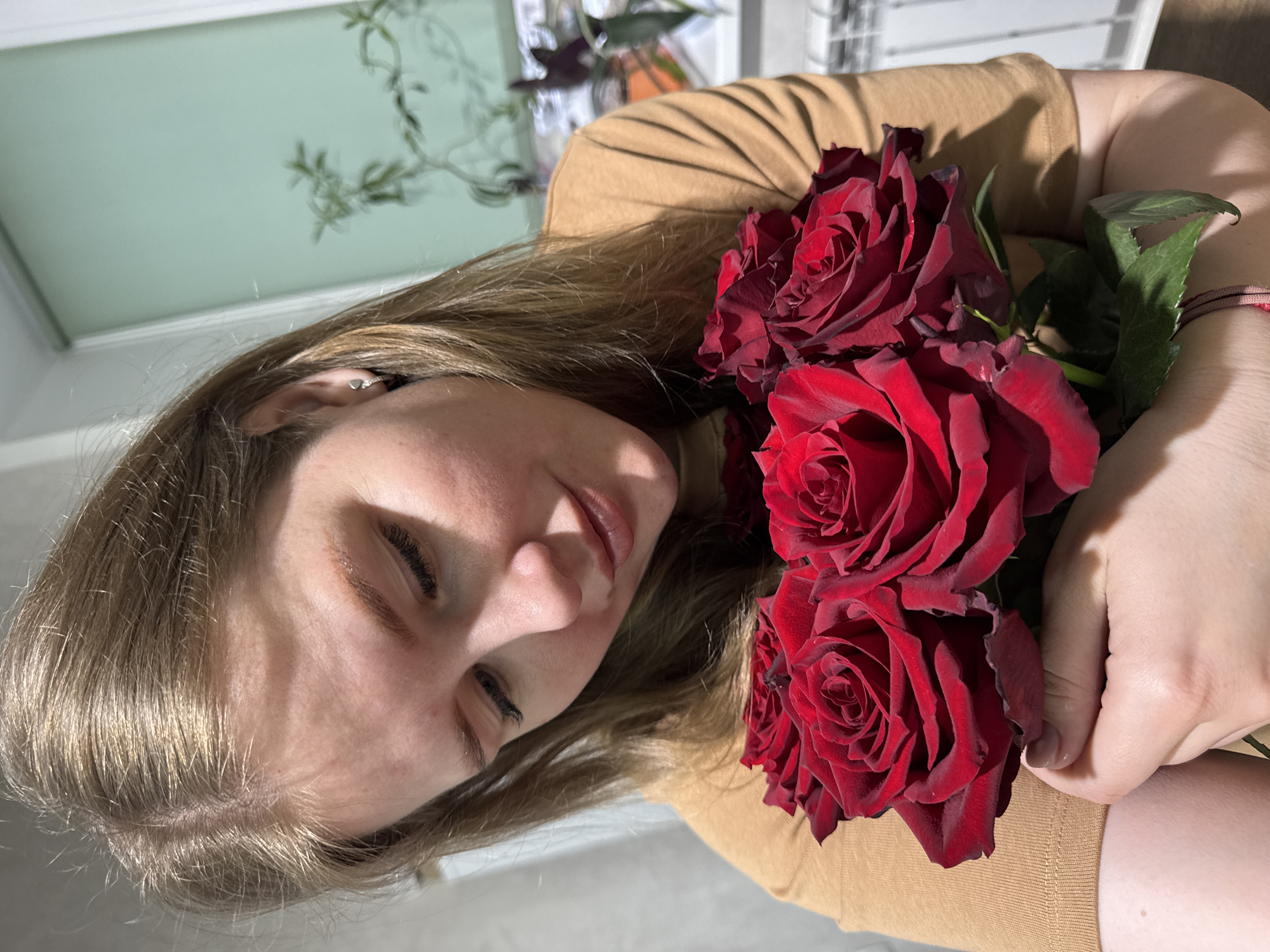 This portrait-style image features a young woman with light brown hair and fair skin, holding a bouquet of three vibrant red roses close to her. She is wearing a light brown, long-sleeved top. Her eyes are gently closed, and a subtle, content smile is visible on her lips, conveying a feeling of serenity, appreciation, or quiet happiness.
A small, simple silver hoop earring is visible in her right ear, and a thin, braided reddish-brown bracelet adorns her left wrist. The roses are prominent, with richly colored red petals and some green leaves visible among them, suggesting they are fresh.
The background is softly blurred, indicating an indoor setting, possibly a home or room. Hints of light-colored walls or a ceiling are discernible, along with indistinct green foliage from what appears to be houseplants or decor. The lighting is soft and even, typical of indoor conditions, without strongly indicating a specific time of day or outdoor weather. No visible text is present in the image. The scene is set in an indoor location in Gornyi (Dvurechenskoe S/P), Russia.