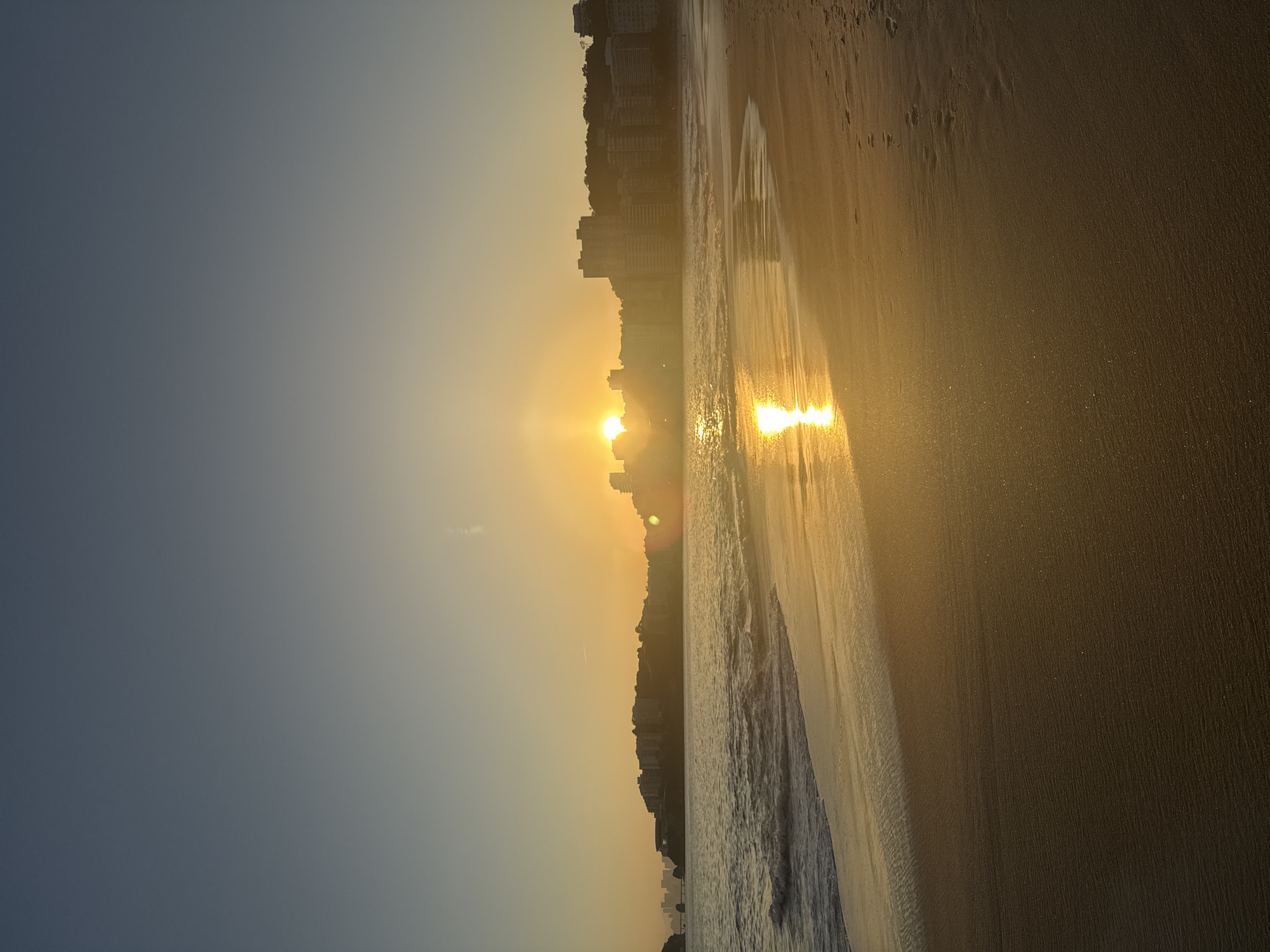 This is a photograph taken at sunset on Icaraí Beach in Niterói, Brazil. The sun is setting over the horizon, casting a warm golden glow across the sky and reflecting on the water. Buildings and hills are visible in the background, silhouetted against the vibrant sunset. The foreground shows the sandy beach with gentle waves lapping at the shore. There are no people clearly visible in the image. The scene is serene and peaceful, capturing the beauty of a coastal sunset.