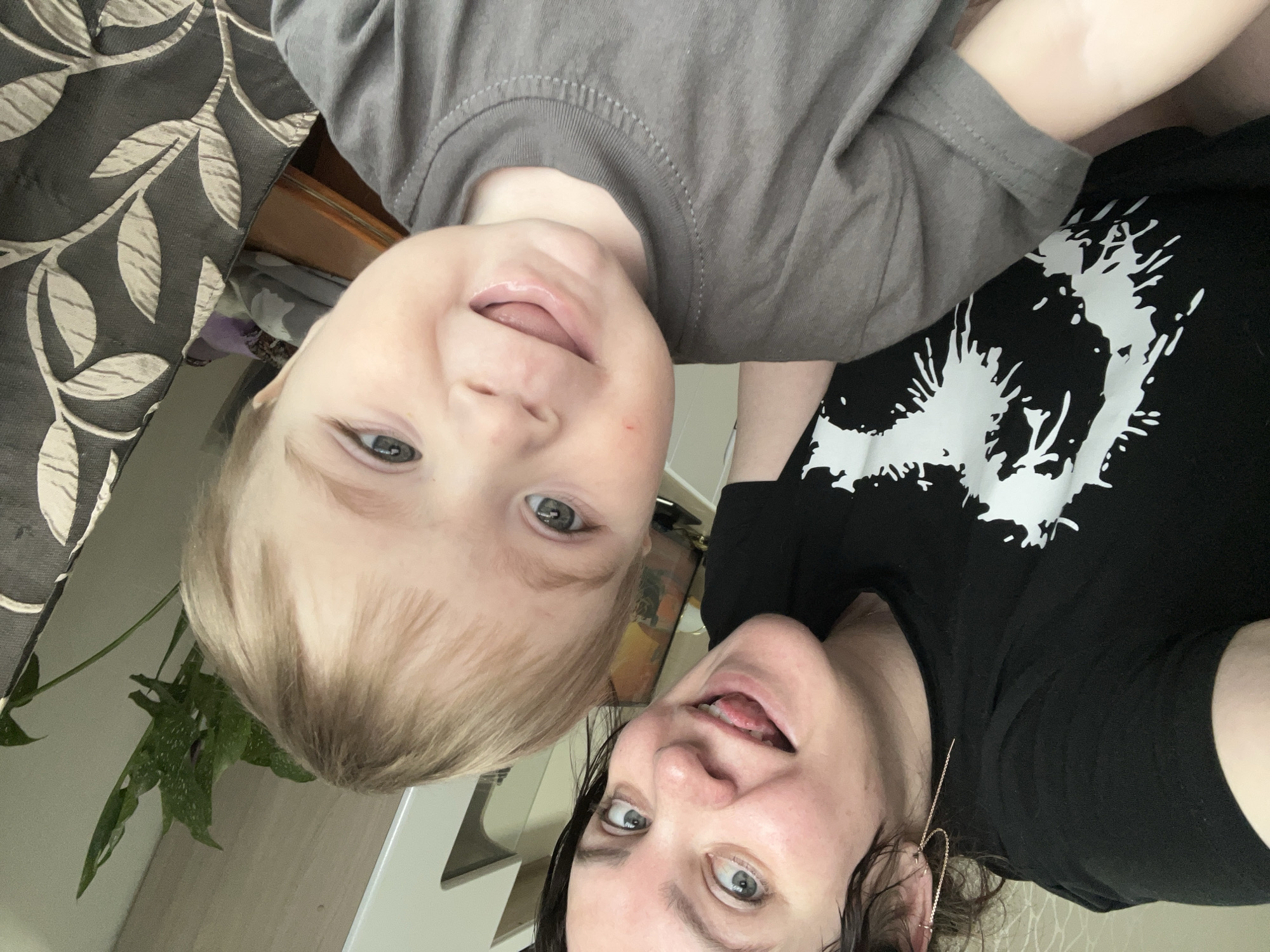 This media file presents a playful, overhead selfie featuring an adult and a young child in an indoor setting.
On the left, a young child with light blonde hair, fair skin, and blue-grey eyes looks directly at the camera with a gentle smile, their tongue slightly visible. They are wearing a plain grey t-shirt. To the right, an adult with dark hair, blue-grey eyes, and fair skin also looks at the camera, playfully sticking out their tongue. They are dressed in a black t-shirt featuring a distinctive white, splattered, or abstract design on the front. A long, gold-colored earring is visible in the adult's right ear.
The background suggests a home environment. To the far left, a dark fabric with a light leaf pattern is partially visible. Below the child's head, green foliage from a plant, possibly a Monstera, can be seen against a light-colored wall. A wooden surface, likely furniture, is also visible behind the child and between the two subjects. A light-toned object is partially visible in the bottom right corner. Both individuals exude happy and playful emotions. There is no visible text in the image. The photo was taken in Perm, Russia.