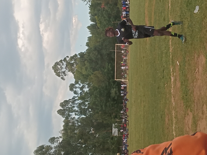 The image depicts an outdoor soccer field in Chevaywa ward, Kenya, under a cloudy sky. In the midground, a male individual with dark skin and braided hair, wearing a black and white soccer uniform (jersey with a green emblem and "MALALA" text, black shorts, black and yellow/green striped socks, and light-colored athletic shoes), runs from right to left across a grassy field. A white line marks the field in the lower-midground. In the background, a white soccer goal is visible. Along the far side of the field, a crowd of spectators is gathered, and behind them, various structures and a dense line of tall green trees extend across the horizon. A partial view of an orange garment is present in the immediate lower-left foreground.