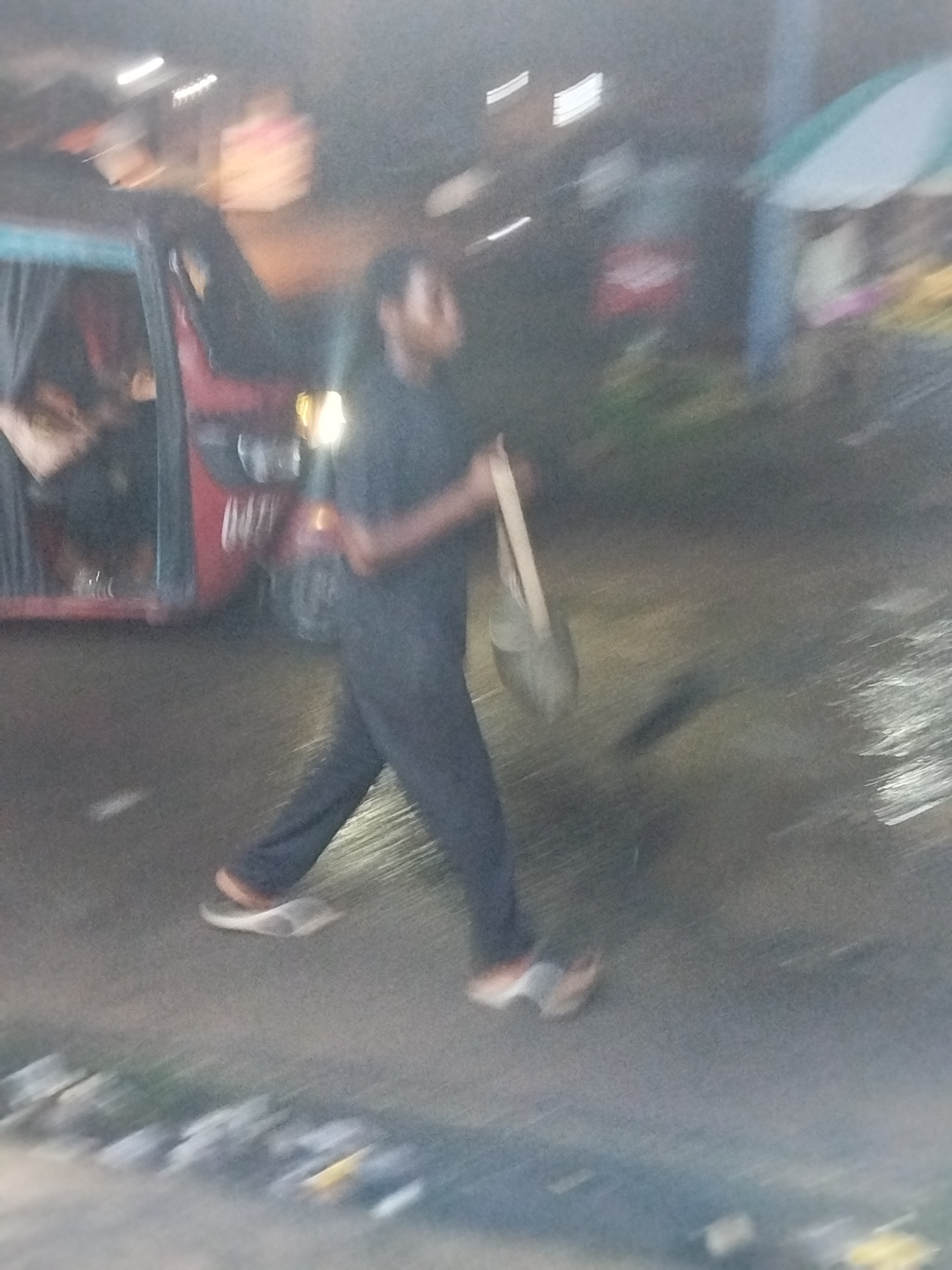 The image shows a man walking on a street in Ikot Mbonde, Nigeria. He is wearing dark clothing and sandals. He is carrying a bag. A red auto rickshaw is parked nearby. There is a food stall with a pale green awning to the right. The scene is blurry, suggesting movement during the capture. There are street lights in the background.