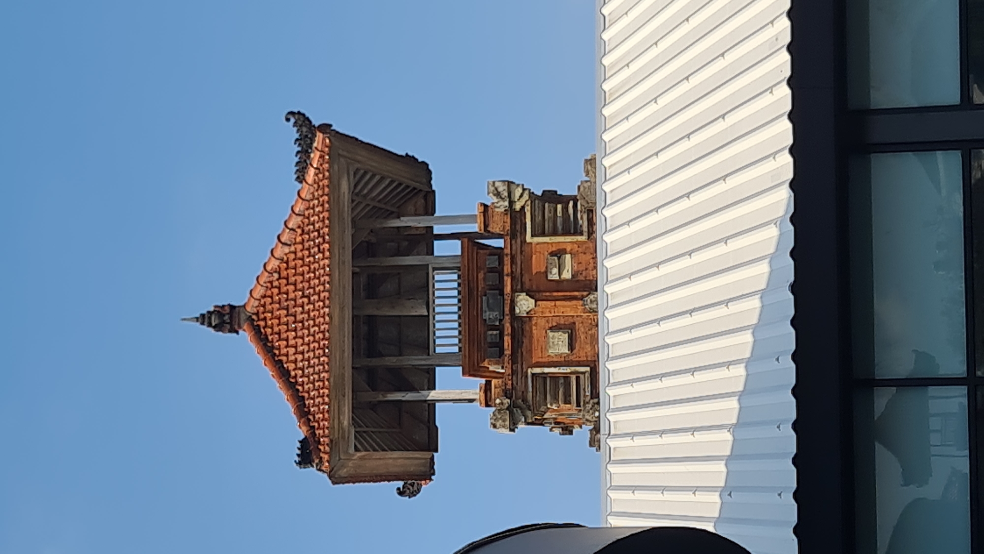 The image shows a traditional Balinese structure with a tiered red-tiled roof, intricate brickwork, and ornate wooden elements, set against a clear blue sky. The structure appears to be a tower or a temple element, possibly part of a larger complex. In the foreground, a modern corrugated metal roof is visible, casting a shadow on a section of it. Below the metal roof, a dark band runs across the image, possibly a facade, with large windows reflecting the sky and the structure. The overall scene suggests an urban environment in Denpasar, Indonesia, where traditional architecture often coexists with modern infrastructure. There are no people or animals visible in the image.
