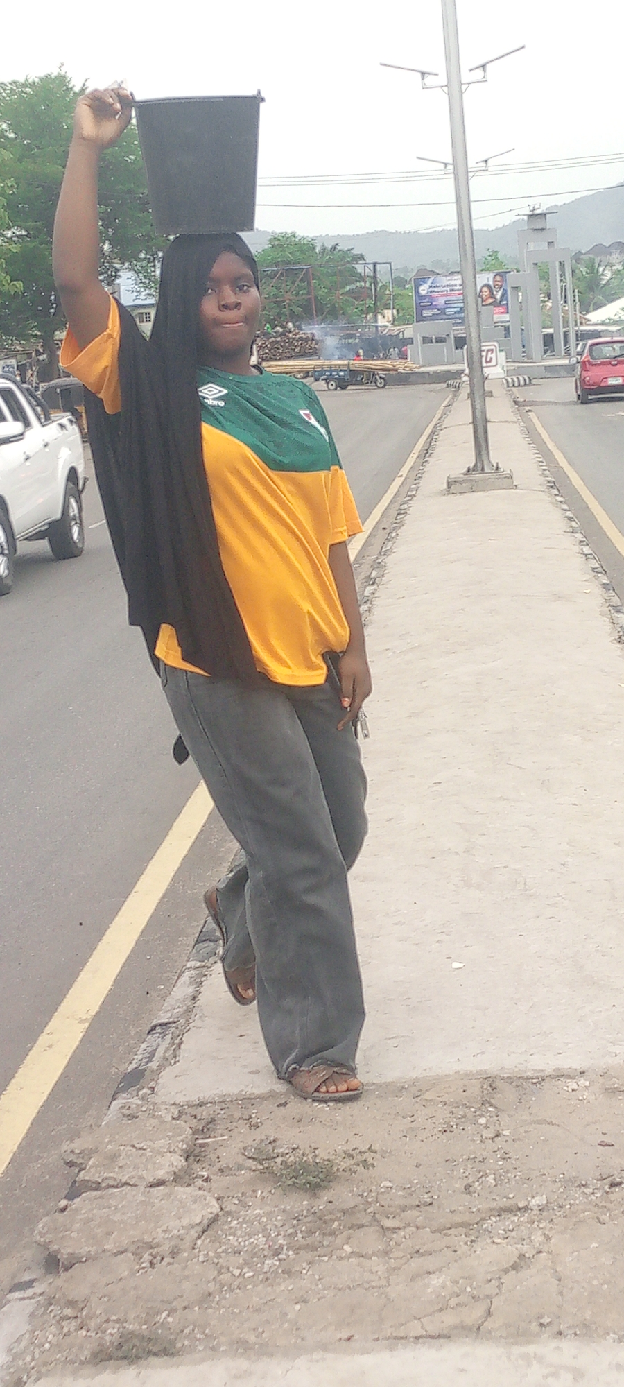 A young woman is balancing a black bucket on her head while standing on a sidewalk next to a road. She is wearing a green and yellow sports jersey, gray pants, and sandals. A black shawl is draped over one side of her body. Cars and traffic signs are visible in the background. Based on visual landmarks, the image appears to be taken in Adankolo, Nigeria. A pile of lumber is on a wagon by the roadside. A white truck is in the lane on the left and a red vehicle in the opposite lane.