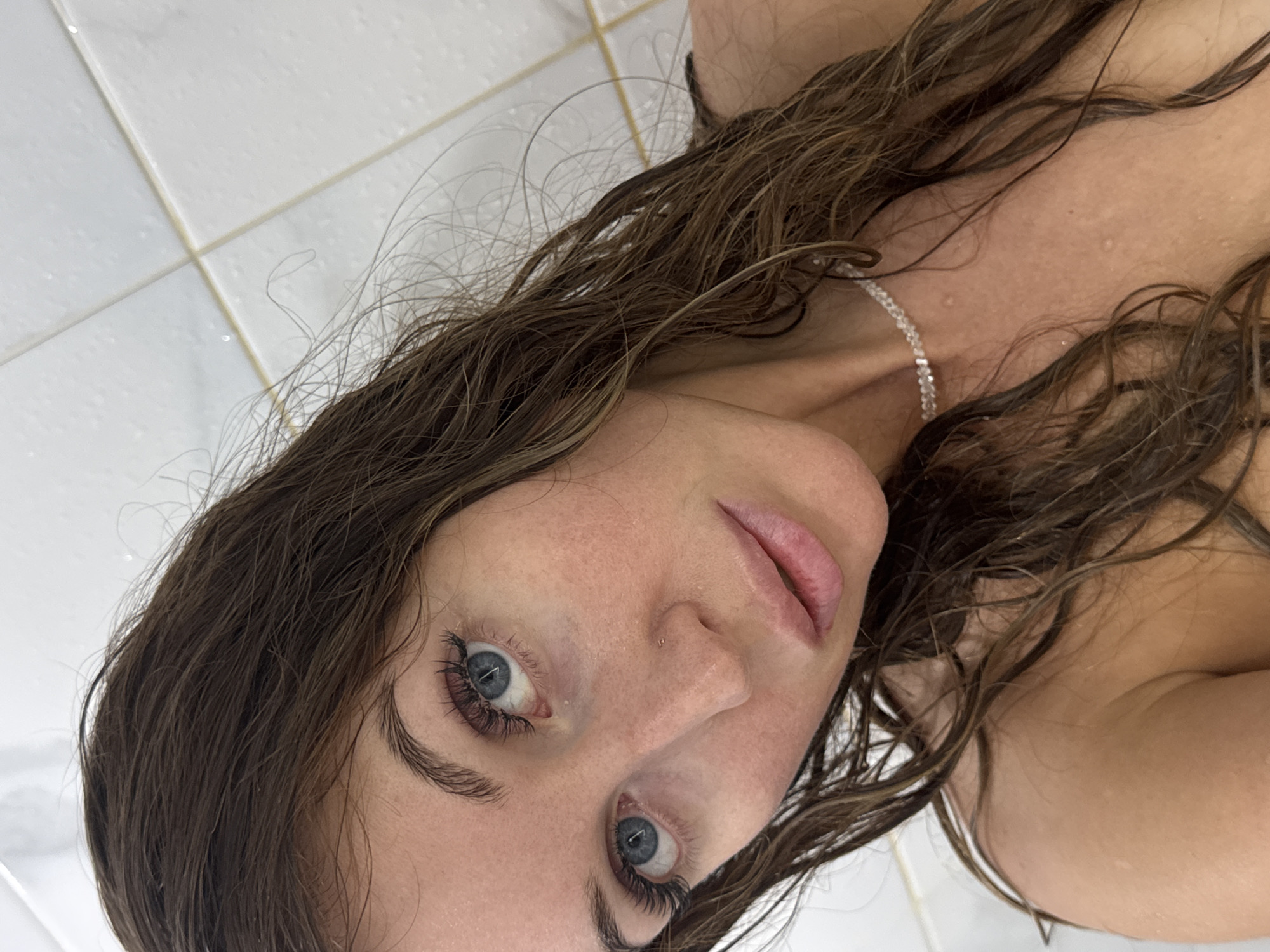 A close-up, angled view of a woman with wet, brown hair. She has fair skin, blue eyes, and full eyelashes. She appears to be wearing a delicate, silver necklace. The background consists of white, tiled wall. The location is not discernible from the provided image.