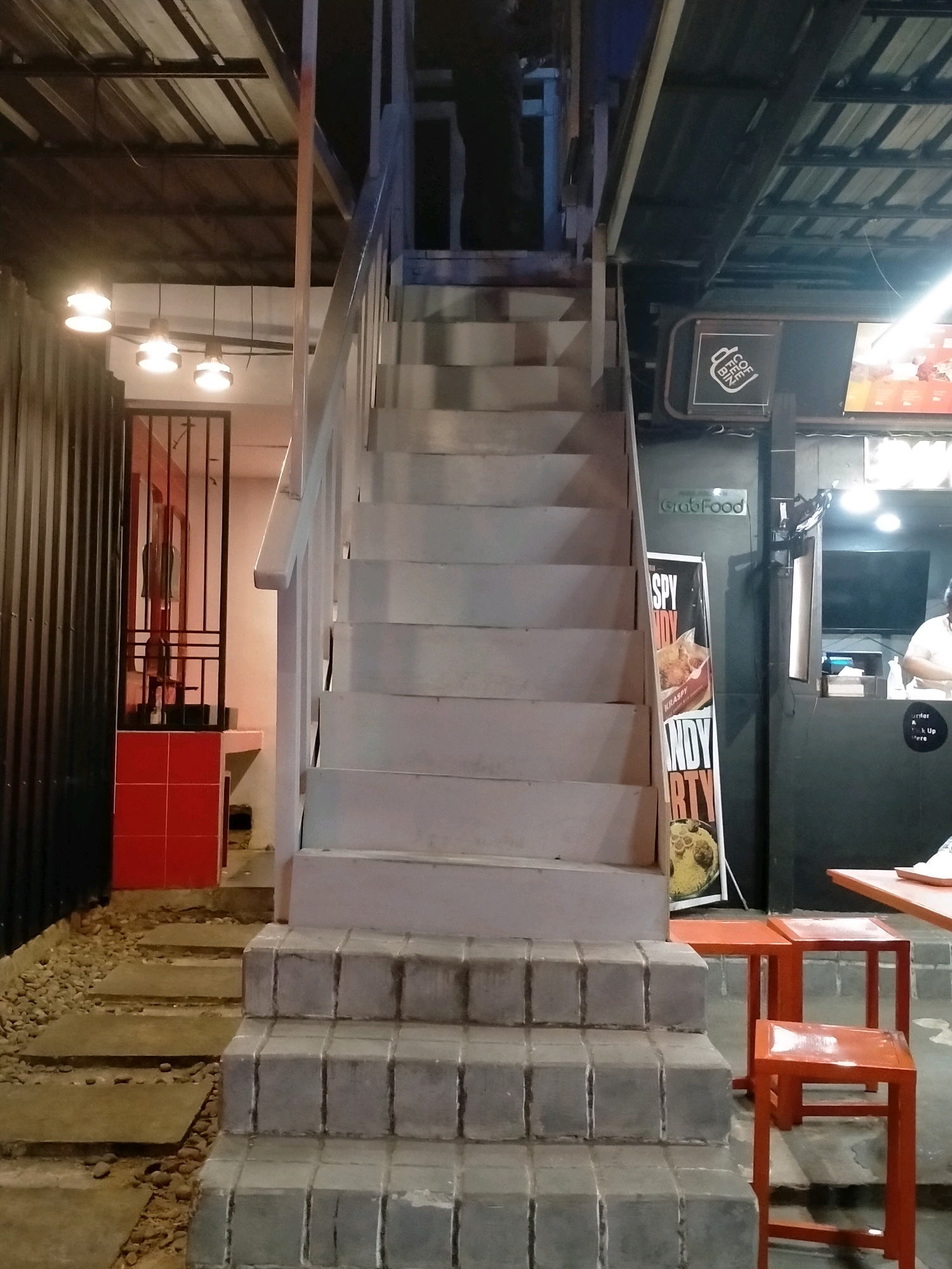 An exterior view of a multi-story building at night in Ambon, Indonesia, features a prominent outdoor staircase made of light-colored concrete blocks with gray tiling. The staircase leads up to an upper level. To the left of the stairs, a path of stepping stones is surrounded by gravel. Further left, a black corrugated metal wall is visible, adjacent to a red-tiled section and an open doorway with a black metal gate leading into a brightly lit room. To the right of the stairs, a food stall is set up with a counter area where a person in a white shirt is visible. An orange promotional stand with text and images is positioned near the stairs. Several small orange stools are placed in front of an orange table. The scene is illuminated by overhead lighting, creating a mix of light and shadow.