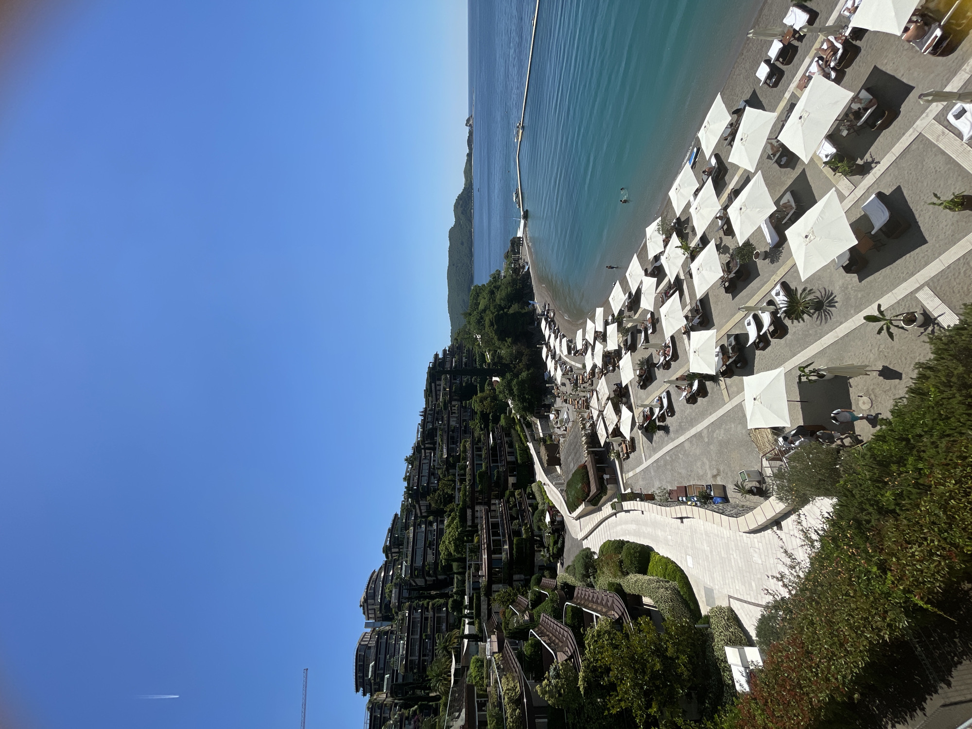 The image presents an elevated view of a coastal scene in Budva, Montenegro, under clear blue skies. The Adriatic Sea borders a sandy beach, where rows of closely packed white umbrellas and lounge chairs are arranged, populated by individuals in beachwear, suggesting leisure activities. Adjacent to the beach is a paved area with manicured gardens and walkways. Several multi-story buildings with green vegetation are terraced up the hillside, which leads to the beach. The area appears to be a resort. In the sky, the contrail of a passing jet is visible. A breakwater extends into the water.