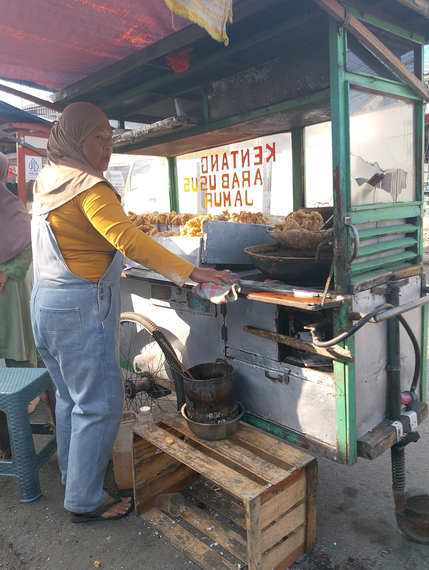 The image depicts a street food stall in Lembang, Indonesia. A woman, wearing a brown hijab and a yellow long-sleeved shirt with blue denim overalls, is standing behind a mobile food cart. The cart has a green metal frame and is filled with food items, including what appears to be fried snacks displayed in baskets and bowls. The cart also has a glass display area with some broken panes. Behind the woman, a sign is visible with orange lettering that reads "KERUPUK UDANG" and "GARAM" in Indonesian, indicating shrimp crackers and salt. To the left of the cart, a blue plastic stool is placed on the ground. A bicycle wheel is attached to the side of the cart. The ground surface is asphalt, with some debris scattered around. The scene is illuminated by natural daylight, casting shadows. In the background, another person in a light purple hijab and green garment is partially visible, and the reflection of a vehicle and buildings can be seen in the cart's glass panels.