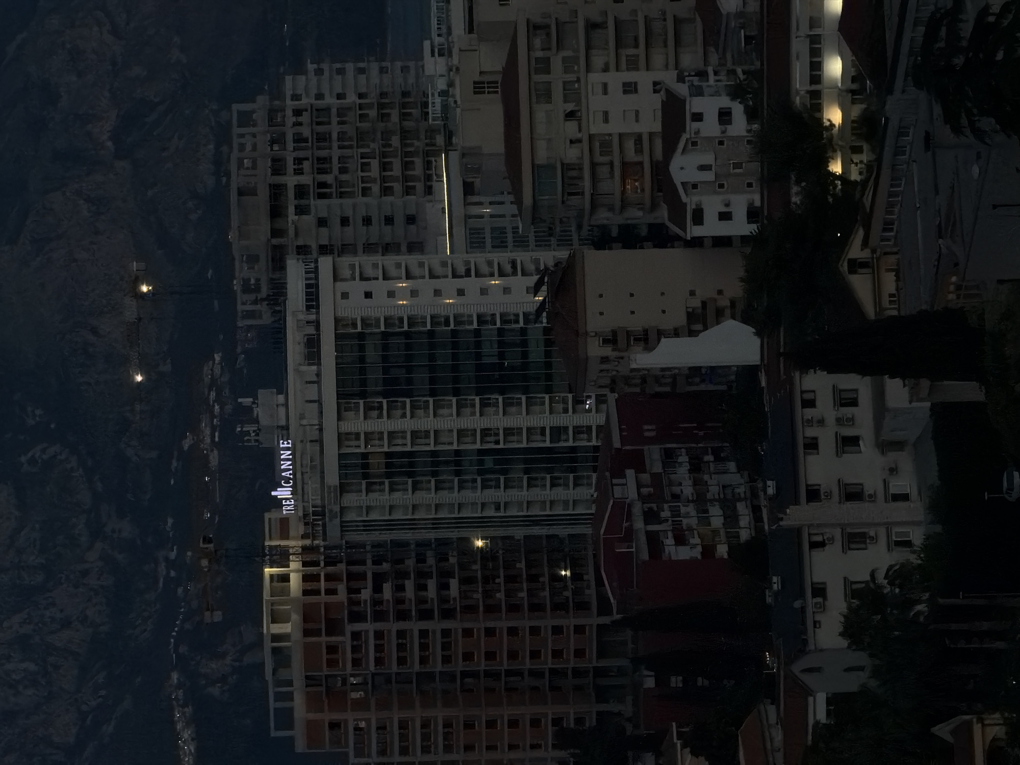 The image shows a nighttime view of Budva, Montenegro. The "TRE CANNE" sign is visible on a high-rise building. Other high-rise buildings are also visible, suggesting a dense urban environment. In the distance, mountains loom over the cityscape. The photo gives a sense of the scale and density of this coastal city at night.
