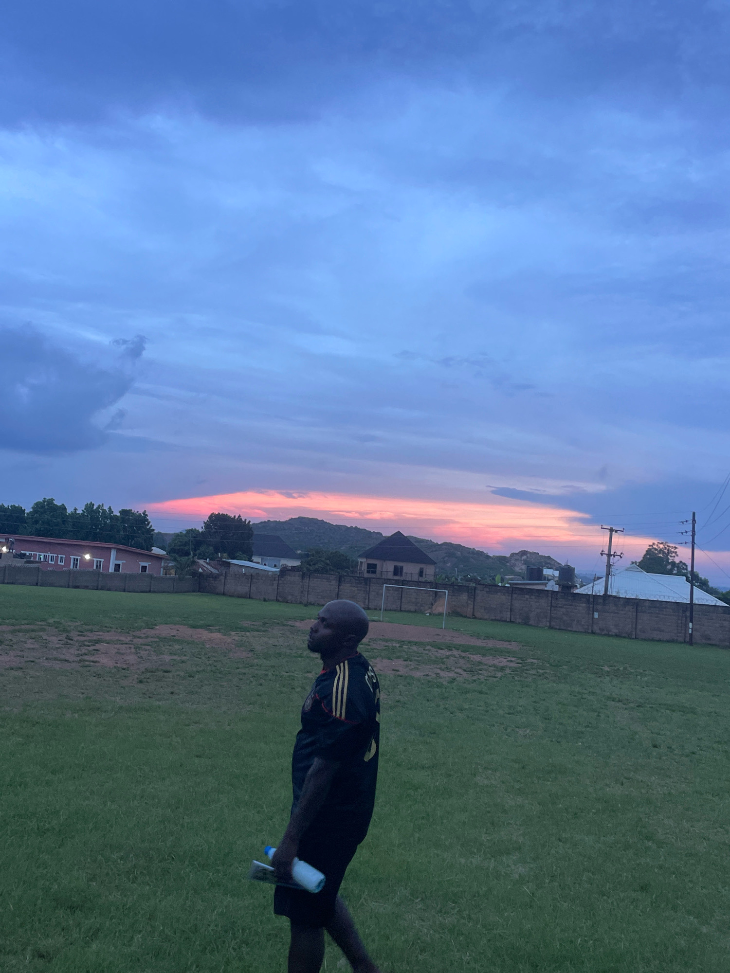 A man wearing a black and yellow striped athletic shirt and black shorts walks across a grassy field. He is holding a clear water bottle and a tablet in his left hand. The field is surrounded by a low brick wall. In the background, there are buildings, trees, and rolling hills under a twilight sky. The sky is a mix of blues and pinks, indicating either sunrise or sunset. The scene is likely in Jos, Nigeria, a city known for its elevated terrain and cooler climate.