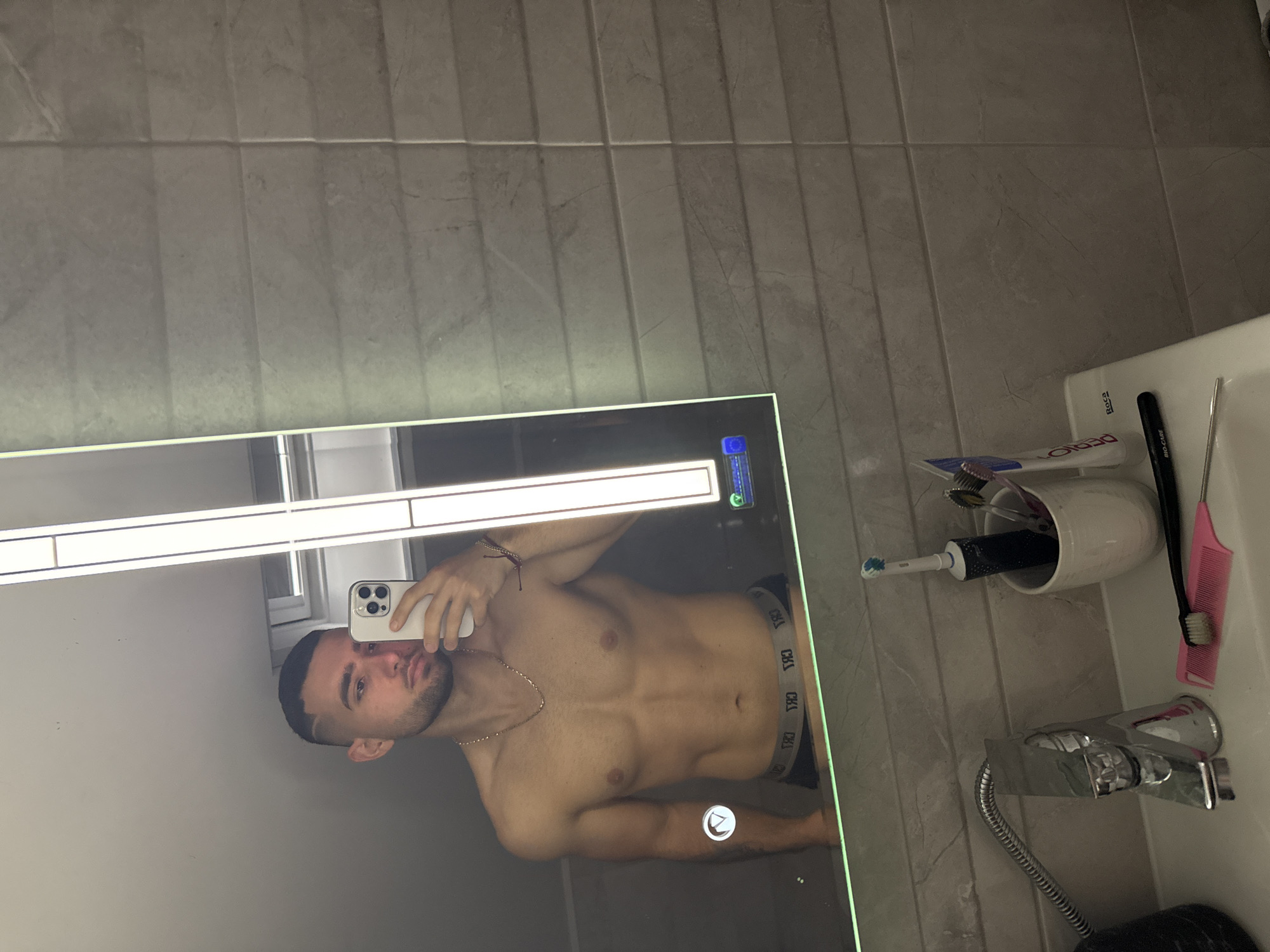 A shirtless man is taking a selfie in a bathroom mirror. The bathroom has gray tiled walls. In the mirror, the man is holding a white iPhone, displaying his face and torso. He has dark hair, a short beard, and is wearing a gold chain. "CK" lettering is visible on the waistband of his underwear. To the right of the mirror is a white sink with a faucet, holding a toothbrush holder containing toothbrushes and a comb. A tube of toothpaste is also present. The location is unknown and no evidence suggests that it is in Gabrovo, Bulgaria.