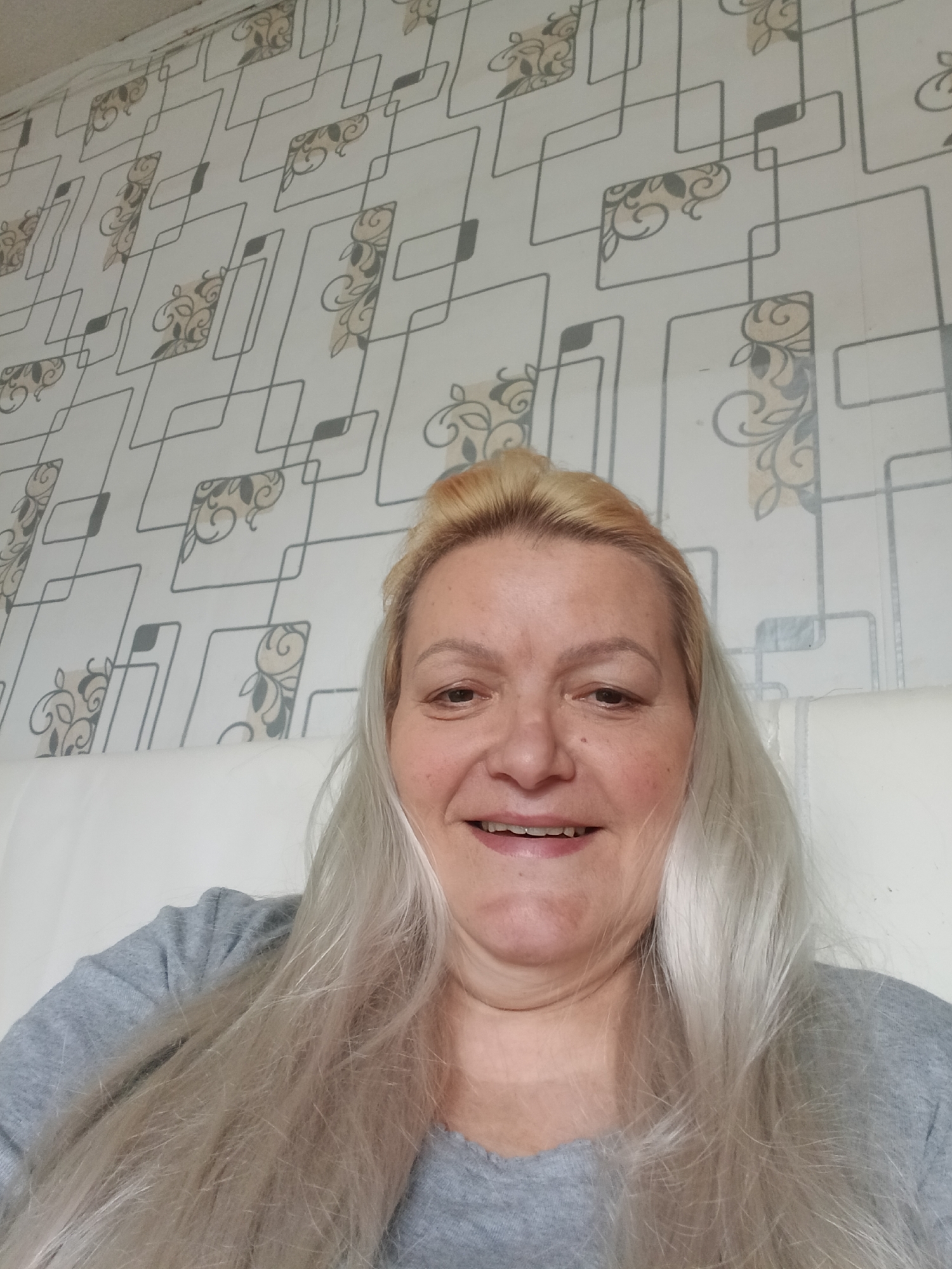 A woman with blonde and grey hair is smiling at the camera. She is wearing a grey t-shirt and appears to be sitting on a white surface, possibly a bed. Behind her is a wall with a geometric patterned wallpaper in white, grey, and tan. The location is unidentified and not confirmed to be Красный Маяк, Russia.