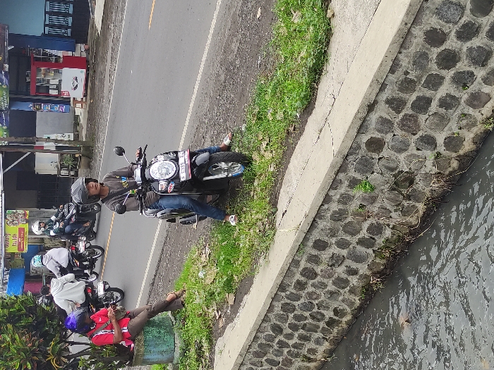 The image, oriented vertically, depicts a bustling roadside scene in Kepanjen, Indonesia, featuring several people and motorcycles alongside a concrete-lined canal.

In the foreground, a man wearing a grey helmet and a long-sleeved grey shirt sits on a black Honda Scoopy scooter, with his feet touching the ground. He is looking towards the left side of the frame. The scooter's license plate, "K 5998 EL," is clearly visible. Behind him, a queue or group of other motorcyclists, also wearing helmets, are stopped on the asphalt road, suggesting they are waiting or parked. One person in a white hoodie can be seen among them. Further to the left, another individual in a red shirt and a purple helmet is seated on a green concrete planter, looking down at an object in their hands, possibly a mobile phone.

The scene includes a narrow asphalt road, a grassy verge, and a concrete-lined drainage canal or ditch containing murky water, which runs along the right side of the frame. On the opposite side of the road, simple commercial buildings or kiosks with awnings and signs are visible, indicating a local business area.

The activity suggests a moment of waiting or resting by the roadside. The time of day appears to be daytime, with soft, diffuse lighting indicating either overcast weather or a time of day without harsh sunlight, as shadows are minimal. Visible text includes the scooter's license plate "K 5998 EL" and partial, indistinct text on background shop signs, possibly including "TOP" and "Kopi" (Indonesian for coffee), and potentially an acronym like "SPBU" (public fuel filling station). The overall setting is characteristic of a local street scene in Indonesia.