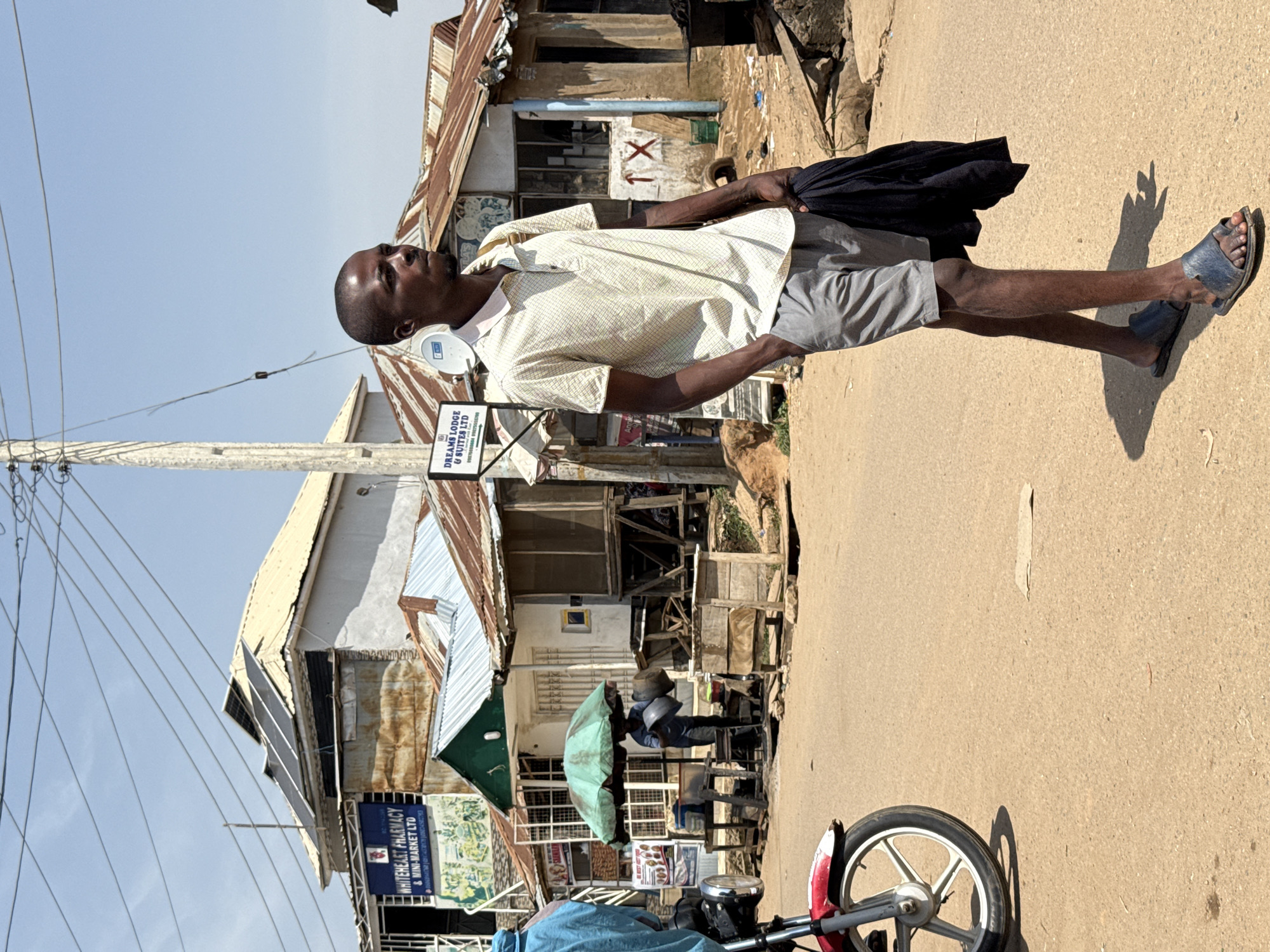 In Abuja, Nigeria, on a bright, sunny day, a young African man walks across a dusty, unpaved street. He is wearing a light-colored, short-sleeved polo shirt with a subtle checkered pattern and grey shorts. He carries a folded dark garment in his right hand and has his left hand in his pocket. He is wearing dark blue sandals.
In the background, a row of buildings with corrugated metal roofs and varying facades line the street. One building, prominently signed "WHITEHEART PHARMACY & MINI-MARKET LTD," has solar panels installed on its roof. Another building is labeled "DREAMS LODGE & SUITES LTD." Several structures appear to be stalls or shops, some with open fronts displaying goods or materials. A green umbrella shades a stall where a person is partially visible, holding two large basins. A red and white motorcycle is parked on the left side of the frame, its front wheel and fender visible. Electrical wires crisscross the sky above the scene, supported by a tall utility pole. The ground is a mixture of dirt and sand, with scattered debris.