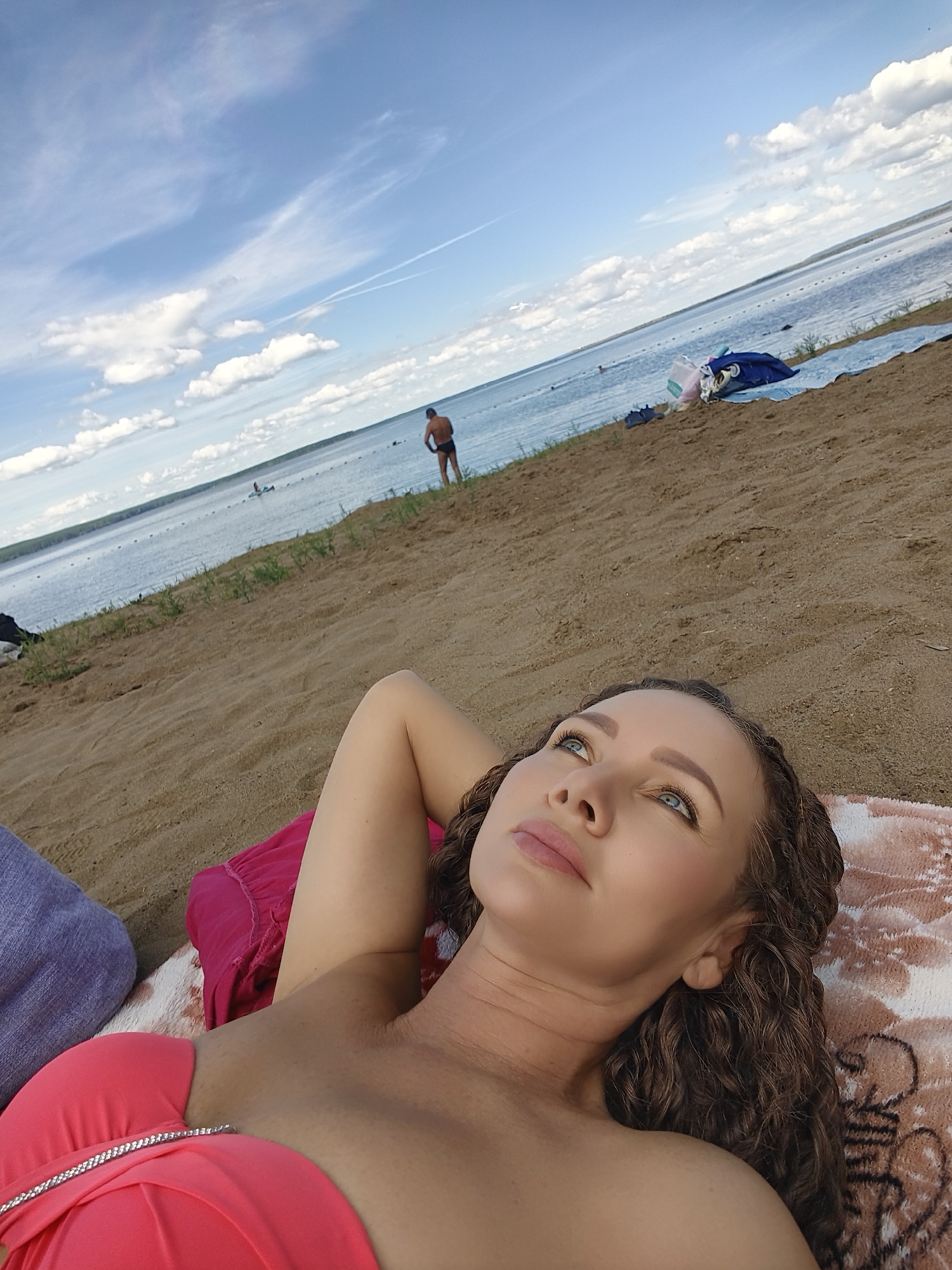 The image shows a woman lying on a sandy beach, looking up at the sky. She is wearing a coral-colored bikini top and has long, curly brown hair. Her head is resting on a patterned beach towel, and her left arm is propped behind her head. Next to her are other beach items, including a blue towel, a blue bag, and a pink cushion.
In the background, a body of water, likely a lake or the sea, stretches out towards the horizon. The water is dotted with people swimming and small buoys creating a boundary. On the far shore, a line of trees is visible. The sky is partly cloudy with fluffy white clouds against a bright blue backdrop. A contrail from an airplane streaks across the sky.
A man in dark swimming trunks is standing at the edge of the water in the mid-ground, his back to the camera. The overall scene suggests a sunny day at the beach, likely in the afternoon given the lighting. The woman's gaze upwards and relaxed posture could indicate a feeling of peace or contemplation. There is no visible text in the image.