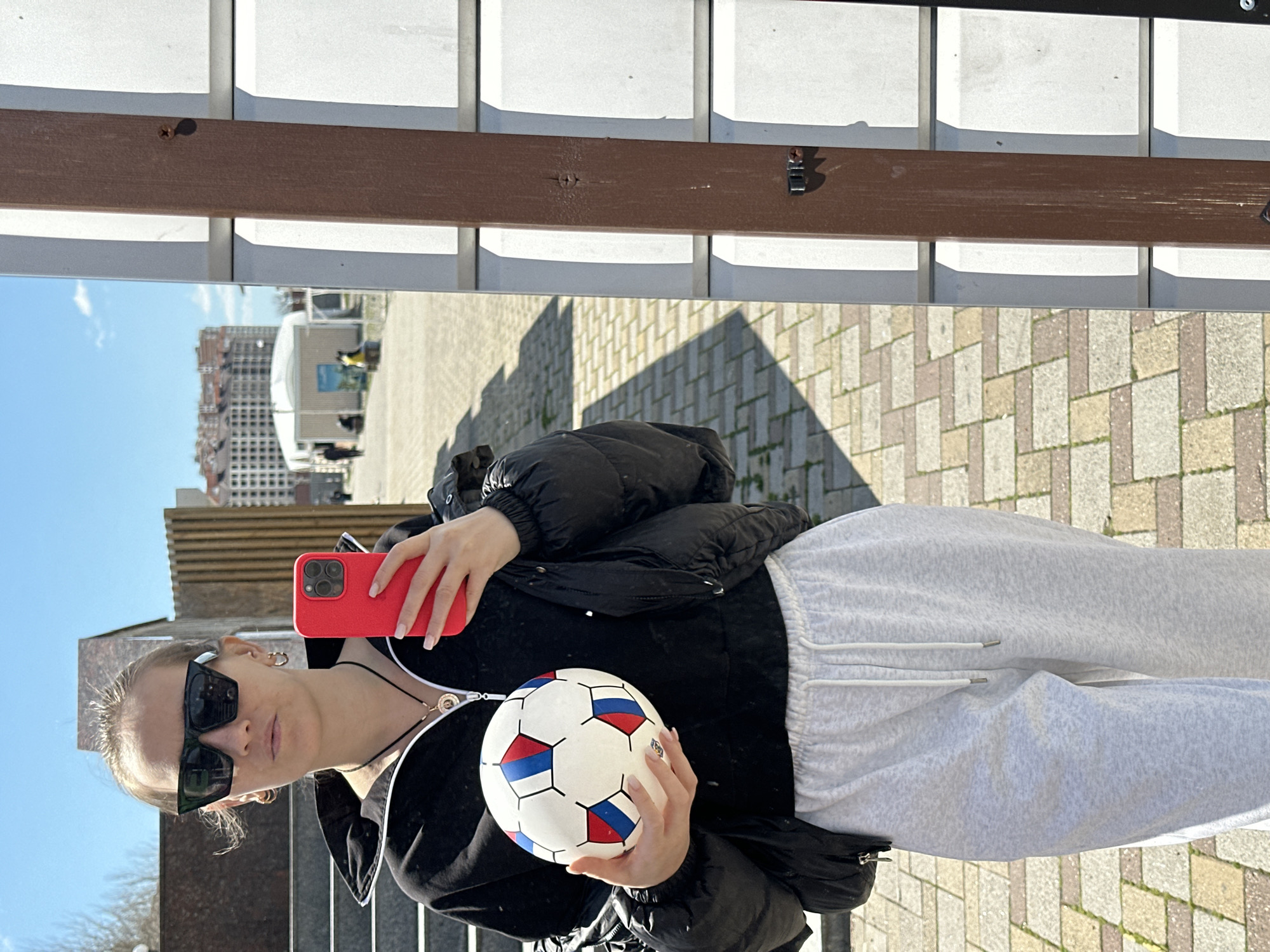 A person wearing sunglasses, a black jacket, and grey sweatpants is holding a red phone in their right hand and a soccer ball in their left hand. The soccer ball is white with red and blue pentagons. The background shows a paved area, possibly a walkway or square, with a light brown and white structure above. In the reflection, buildings can be seen under a clear blue sky. The location is Myskhako, Russia.