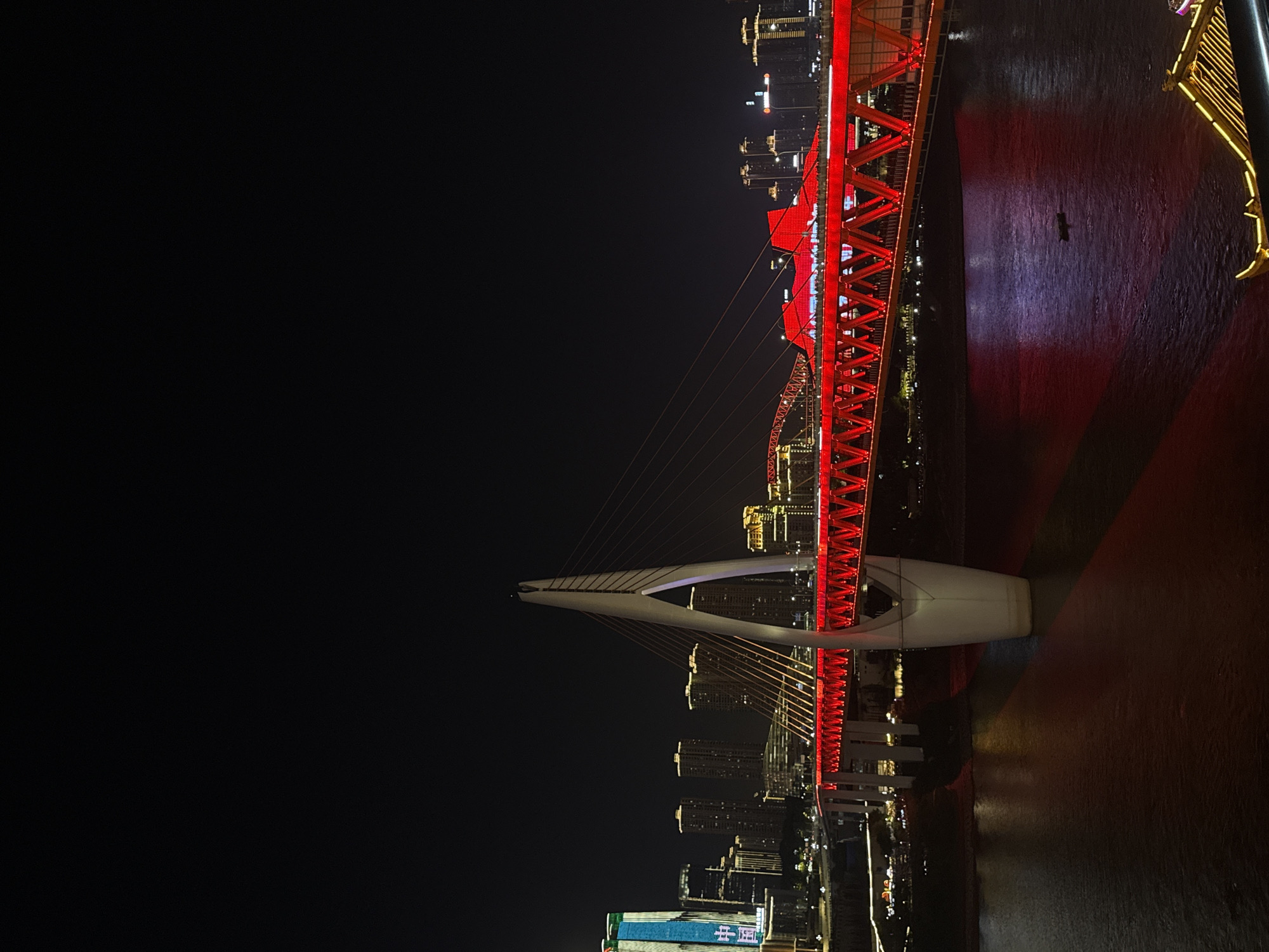 This nighttime photograph shows the Chongqing Dongshuimen Bridge, a landmark in the Yuzhong District of Chongqing, China. The bridge features a prominent red steel truss structure, brightly illuminated, and a white, curved support tower. The bridge spans a dark body of water, reflecting the bridge's red light. The skyline of Yuzhong District is visible on both sides of the bridge, showcasing illuminated buildings and skyscrapers. The background is largely dark, emphasizing the illuminated bridge and cityscape.