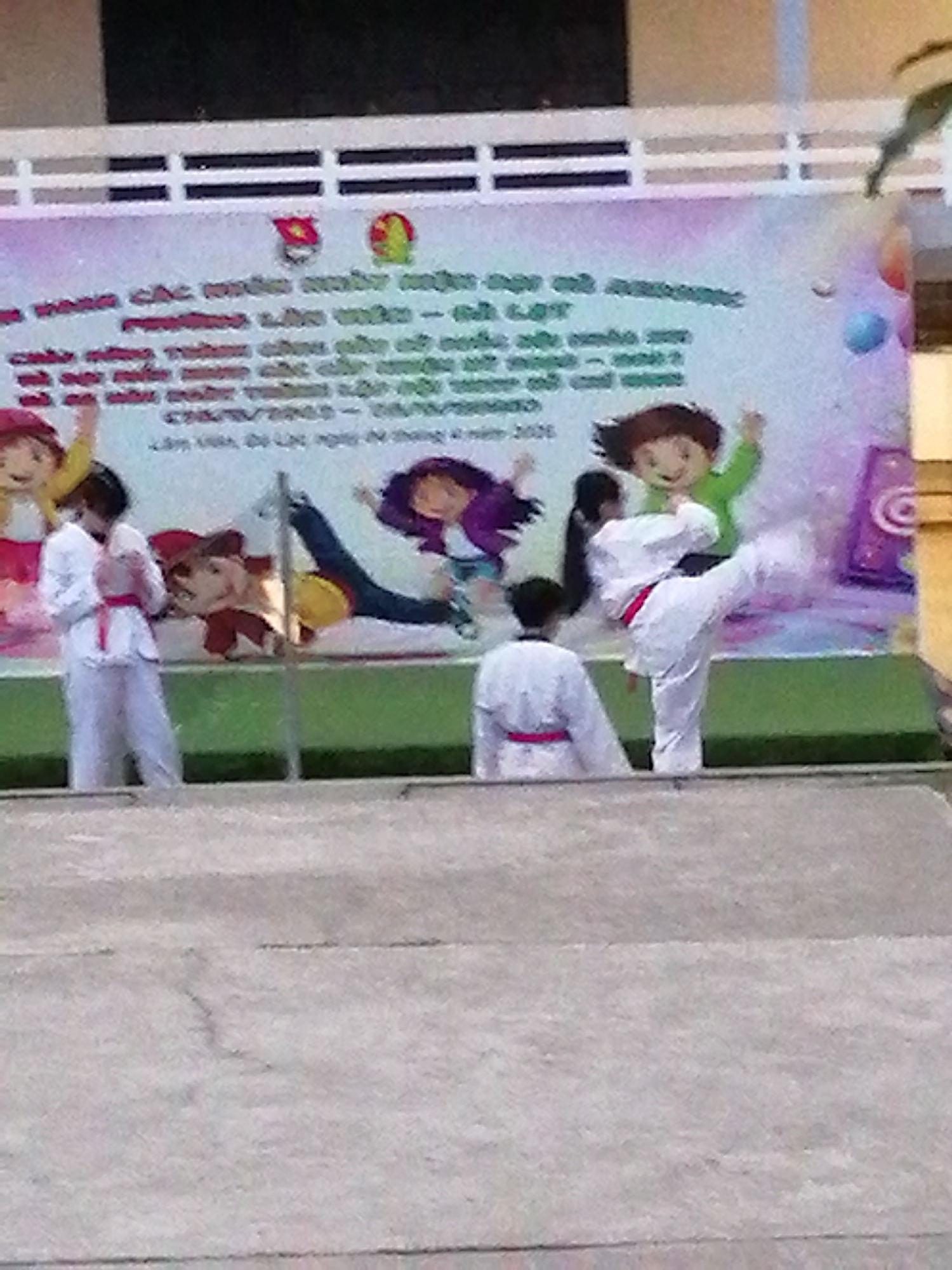 In Lâm Viên - Đà Lạt, Vietnam, a group of children in martial arts uniforms are participating in an event. A young boy in a white uniform with a red belt is captured mid-kick. To his left, a girl in a white uniform with a red belt stands facing away from the camera. Another young boy in a white uniform with a red belt is visible in the background. Behind the children, a banner with colorful cartoon characters and Vietnamese text is displayed. The banner appears to be a backdrop for the event, which is likely a martial arts demonstration or competition. The ground in front of the stage is a light-colored concrete surface.
