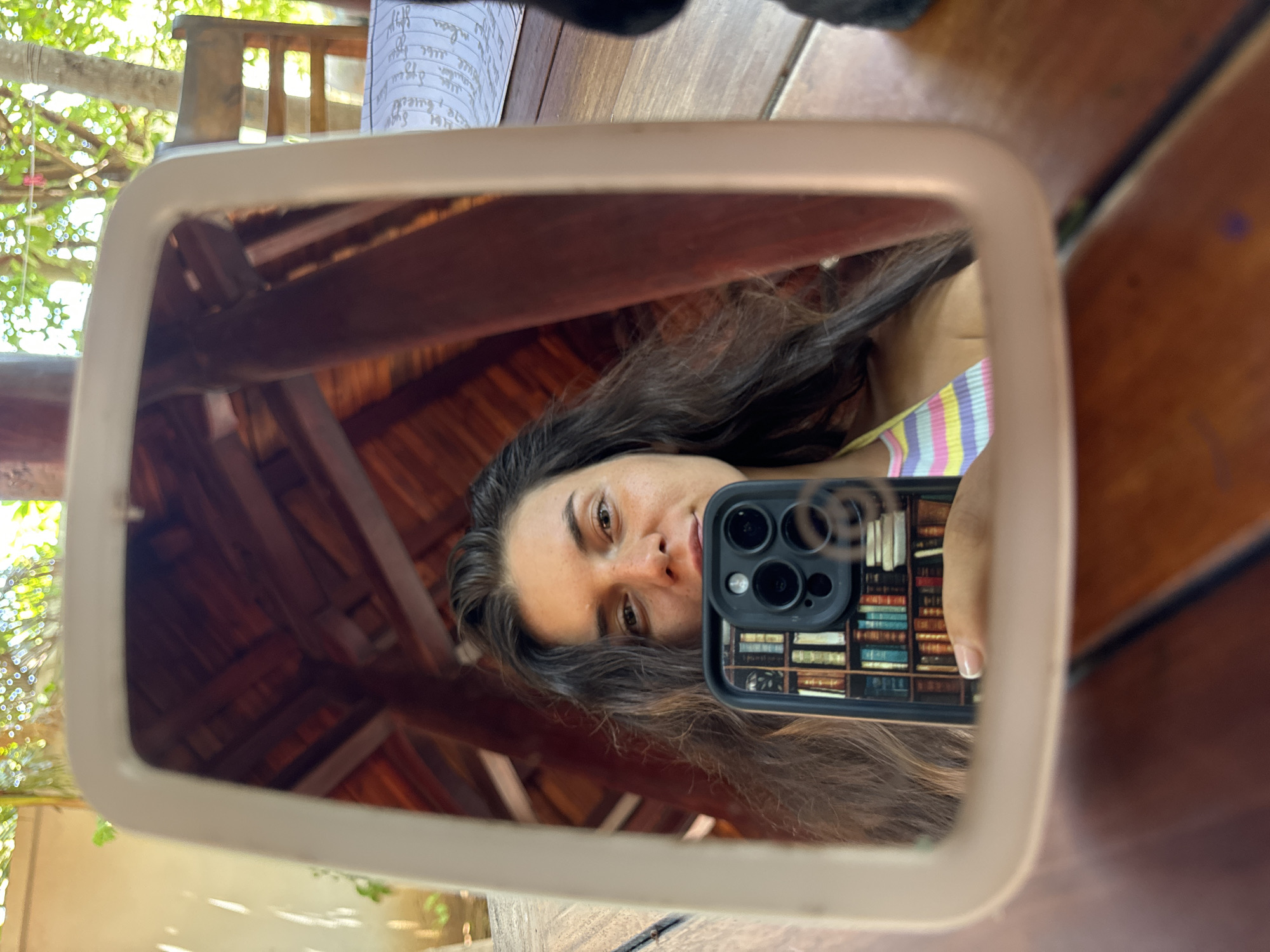 The image shows a woman taking a selfie in a mirror in Mũi Né, Vietnam. She is holding a smartphone in front of her face, and the phone's case has a bookshelf design. The woman has long, dark hair and is wearing a striped bikini top. In the reflection of the mirror, we can see a wooden roof structure and some greenery. The setting appears to be outdoors, possibly at a restaurant or resort, given the wooden table and chairs visible.