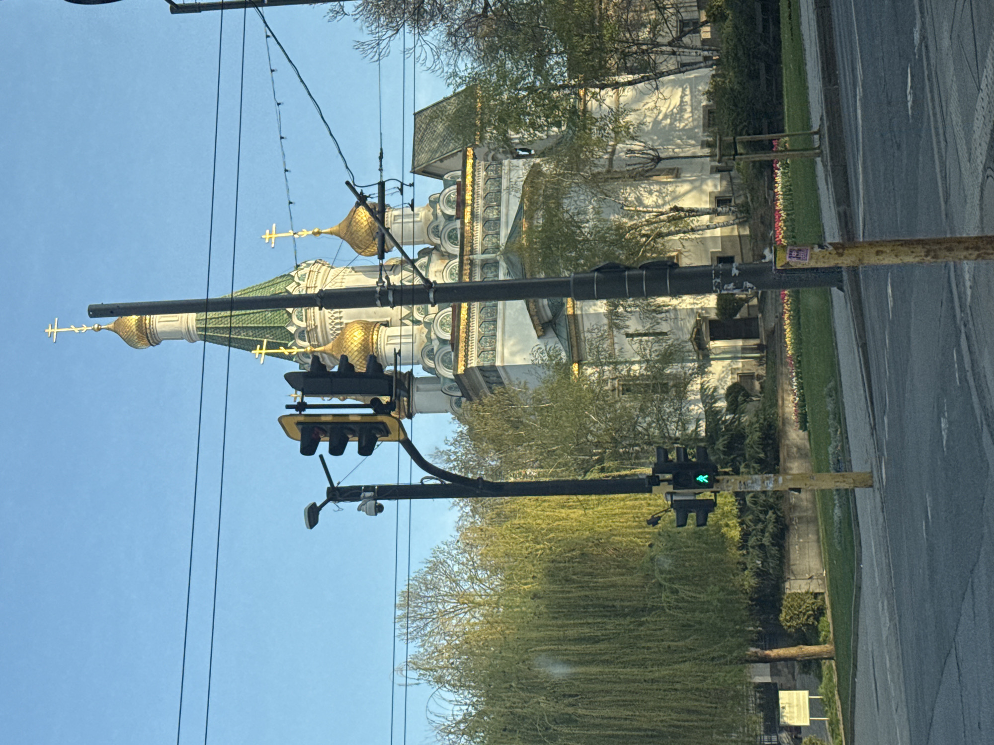 This image, taken in Sofia, Bulgaria, captures a vibrant street scene with the prominent Russian Church (Church of St. Nicholas the Wonderworker) in the background. The church features distinctive golden onion domes, each topped with an ornate cross, and green tiled roofs. Its light-colored facade is richly detailed with intricate white and gold architectural elements, including arches and decorative columns.
In the foreground, a paved street runs alongside a grassy median strip adorned with trees, some with delicate spring leaves, and a colorful bed of tulips. Overhead, a series of dark wires crisscrosses a clear, bright blue sky. Two black traffic light poles are visible; the one closer to the viewer displays an illuminated green left-turn arrow, indicating active traffic flow. Another traffic light further down the street also shows a green signal. A weathered yellow pole, possibly a utility pole or signpost, stands near the street with some faded, unreadable text or posters attached. The scene depicts a clear, bright daytime, suggesting fair weather. No people are visible in the frame, but the urban setting implies a populated area.
