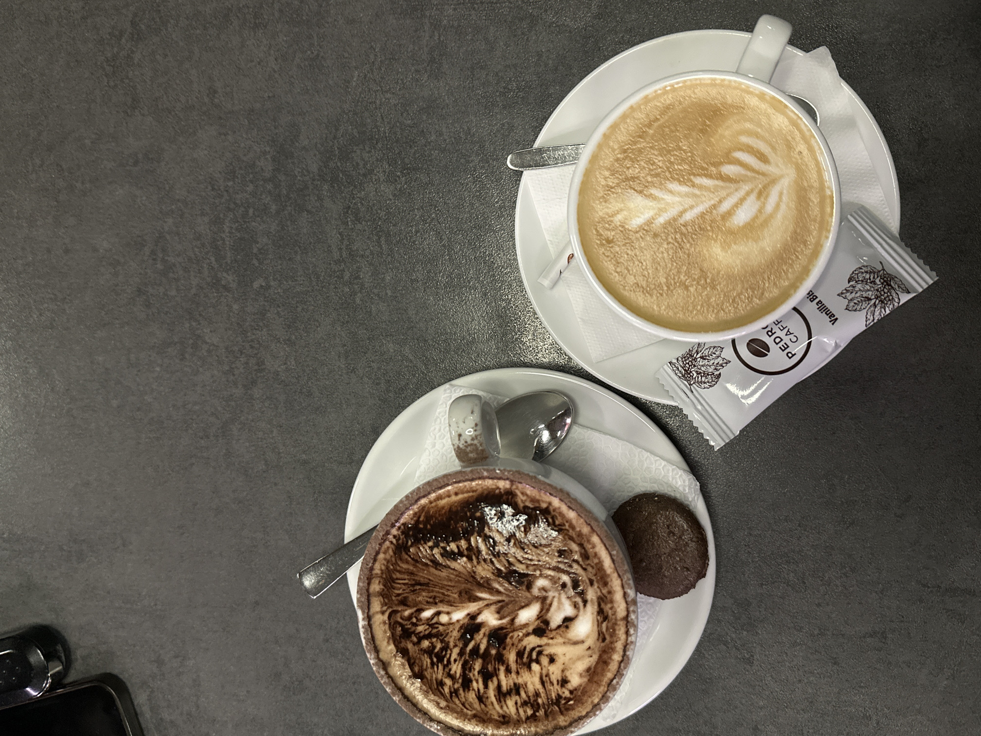 The image shows a top-down view of two cups of coffee on a grey table, likely in Stara Zagora, Bulgaria based on the sugar packet brand "Pedro Caffe" Vanilla Biscuit. One cup contains a latte with latte art depicting a leaf pattern. Next to this cup is a small, rectangular, white sugar packet. The other cup contains a dark, possibly mocha, drink with intricate swirl patterns in the foam. A small, round chocolate cookie is near the mocha cup. Both cups are sitting on white saucers, each with a spoon. In the lower left corner, a small black object is visible. The table has a uniform, matte surface.