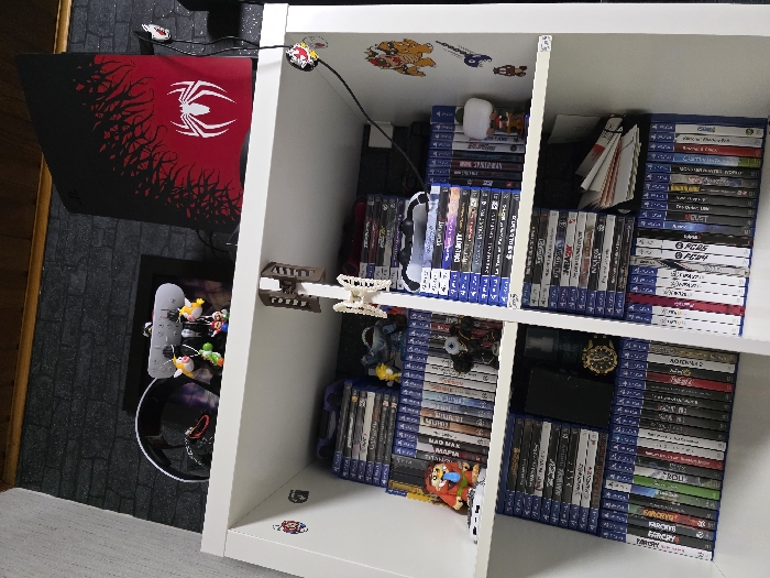 The scene depicts a multi-tiered white shelving unit situated indoors against a dark grey brick-patterned wallpaper, beneath a wooden ceiling trim. The location is an unidentifiable room within the city of Zweibrücken, Germany.
On the uppermost shelf, a black and red PlayStation 5 console, customized with a white Spider-Man logo and red web-like graphics, stands vertically on the right. To its left, a white accessory stand or charging station holds multiple small character keychains, including representations of Yoshi and Mario. A black gaming headset is positioned behind this stand, and a power strip is mounted on the underside of the shelf above it. A black cable with a red and black circular clip descends from the shelf's right edge.
The second shelf displays numerous horizontally stacked PlayStation 4 game cases. A red and orange character figurine with an extended tongue is placed on the left stack, alongside a dark, multi-eyed monster-like figurine. On the right side of this shelf, a white PlayStation controller rests on game cases, and a small Pop! Vinyl-style figurine is present. The vertical wall segment to the right of this shelf features several decals, including a Bowser character and other smaller graphics.
The third shelf also contains horizontally stacked PlayStation 4 game cases. A black smartphone lies flat on the cases to the left, next to a gold-colored wristwatch and a blue glass bottle, possibly containing fragrance. The right section of this shelf holds additional game cases and a stack of white papers or booklets.
The lowest visible shelf is densely packed with PlayStation 4 game cases arranged vertically, showcasing their blue spines across the entire width of the unit. No individuals are present in the image.