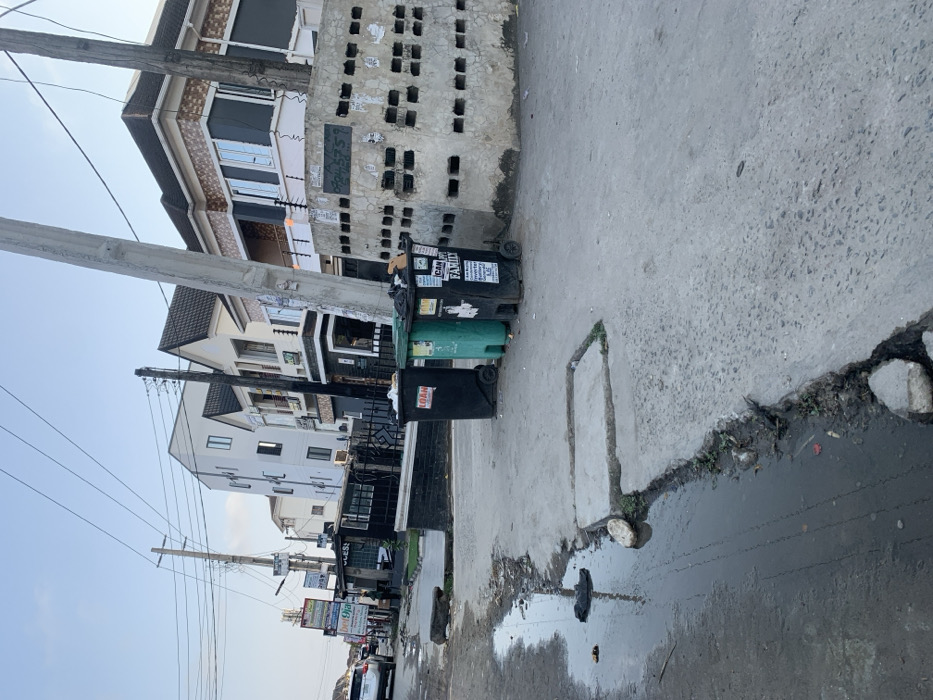 The image displays an urban street scene in Ikota, Nigeria, under clear daylight conditions. The foreground presents a broken concrete road surface with a significant, elongated puddle of dark liquid containing debris, including a dark plastic bag and other refuse, on the left. A raised concrete section, possibly a drain cover or part of a damaged sidewalk, is visible adjacent to the puddle.
Towards the center, three wheeled refuse bins stand on the concrete roadside: two black and one teal-green. The black bin on the far left has the word "LOAN" in red. The black bin on the far right has "ICAN FAMILY" and other text visible. All bins appear partially full or overflowing. Immediately behind these bins and extending to the right is a low, weathered concrete block wall featuring multiple rectangular openings arranged in rows. This wall has various posters and graffiti affixed to its surface.
In the midground, a series of multi-story buildings line the street. These structures exhibit modern architecture with varying facades, including white stucco, dark grey panels, and patterned brick/tile accents, typically featuring multiple windows and balconies with dark railings, some equipped with electric or barbed wire security features.
On the left side of the street, further down, utility poles heavily laden with numerous overhead wires are visible, along with multiple commercial signboards. One sign clearly displays "bet9ja," and another "ACCESS." A small structure, possibly a kiosk, stands near these signs. A single individual, indistinctly visible, stands near this kiosk. A white SUV is parked or moving on the road in the distance on the left. A prominent, light-colored utility pole stands vertically in the right foreground, partially obscuring the view of the buildings beyond. The sky above is light blue and clear.