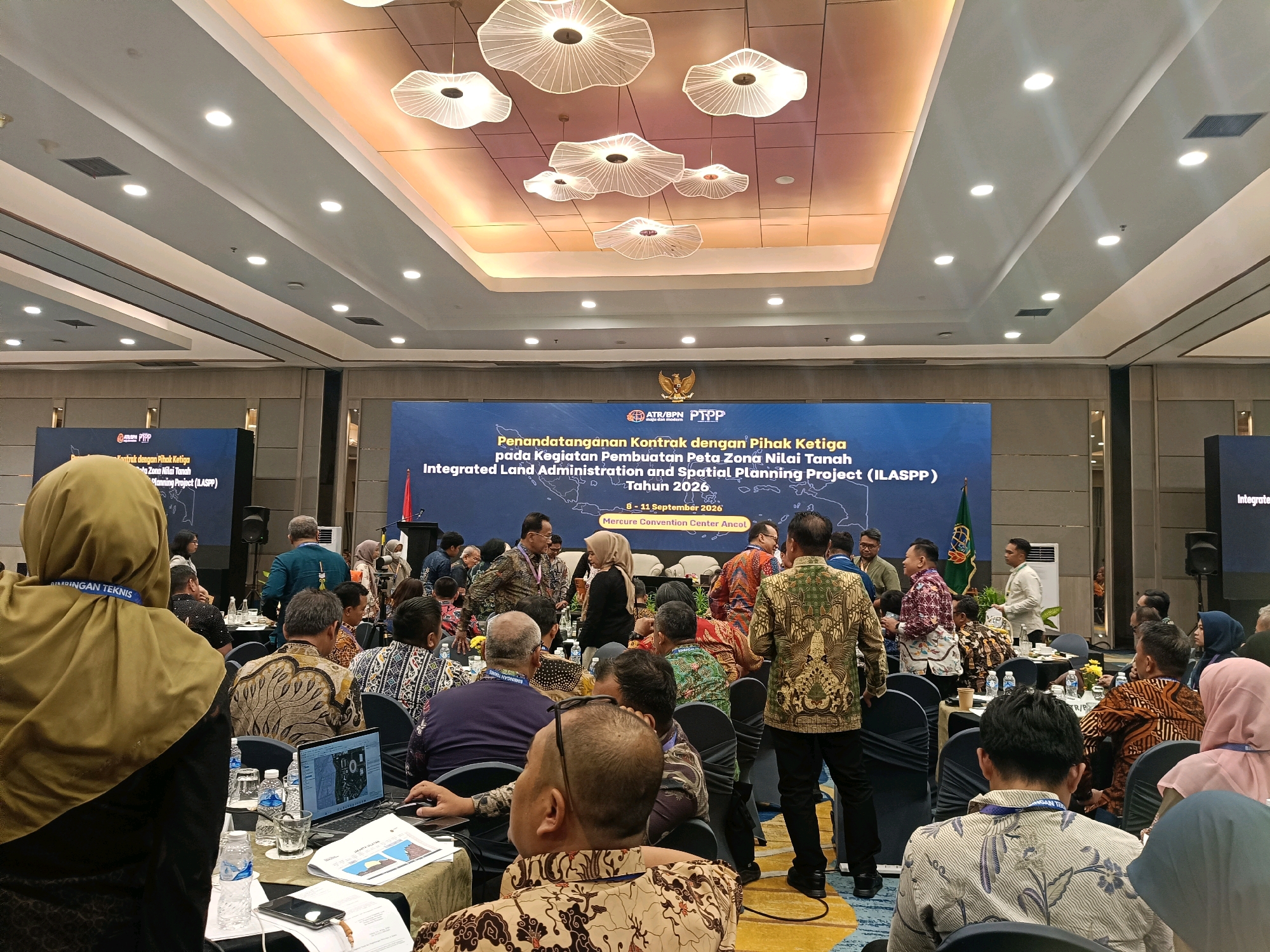 The image depicts an indoor event, likely a conference or seminar, taking place in a large ballroom. The venue has a high ceiling with modern, decorative lighting fixtures and recessed lighting. A large screen in the background displays text in Indonesian and English, indicating the event is "Penandatanganan Kontrak dengan Pihak Ketiga pada Kegiatan Pembuatan Peta Zona Nilai Tanah Integrated Land Administration and Spatial Planning Project (ILASPP) Tahun 2026," which translates to "Signing of Third Party Contracts for the Land Value Zone Map Creation Activities Integrated Land Administration and Spatial Planning Project (ILASPP) Year 2026." Below this, it states "8-11 September 2026" and "Mercure Convention Center Ancol." This confirms the location as the Mercure Convention Center in Ancol, North Jakarta, Indonesia.
A diverse group of people, predominantly adults, are seated at round tables and standing in the aisles. Many attendees are wearing batik shirts, suggesting a cultural element to the event. Some individuals are engaged in conversation, while others appear to be listening to presentations or looking at materials on their tables. Laptops, water bottles, glasses, and documents are visible on the tables. In the foreground, a woman wearing a hijab is facing away from the camera, partially obscuring the view of the attendees at her table. The overall atmosphere appears to be professional and formal.