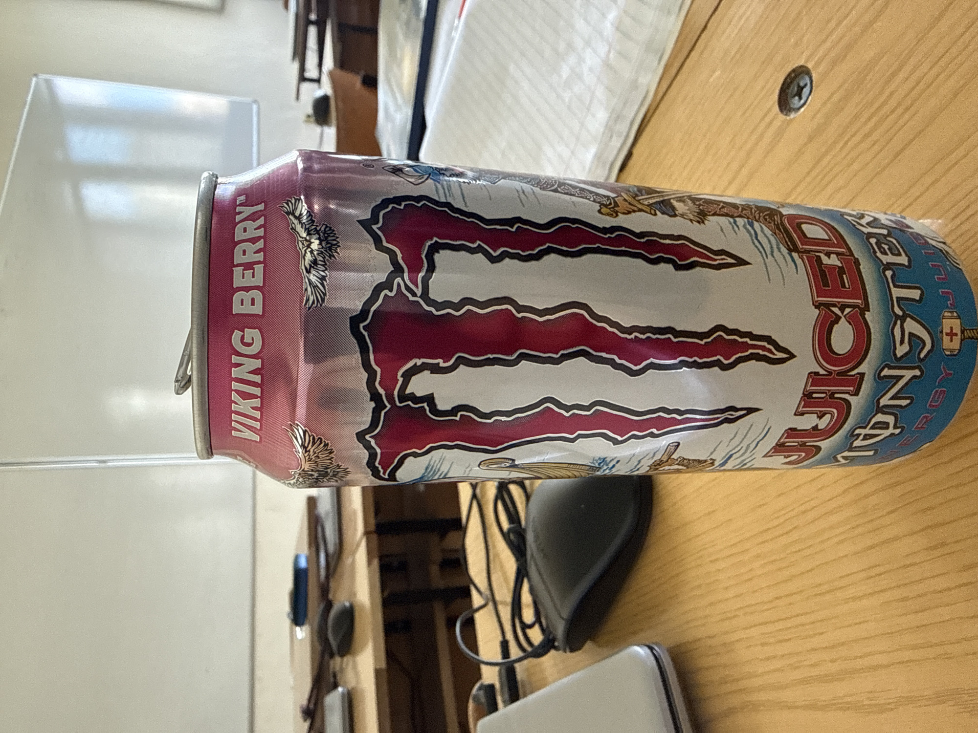 The image shows a can of Monster Energy Juiced Viking Berry drink lying on a wooden table. The can is positioned horizontally in the center of the frame. To the left, a whiteboard is visible in the background. A black computer mouse and a portion of a white laptop are situated near the can. The setting appears to be an indoor environment, possibly an office or study area. There is no evidence to indicate the location is Varna, Bulgaria. No people are present in the image.