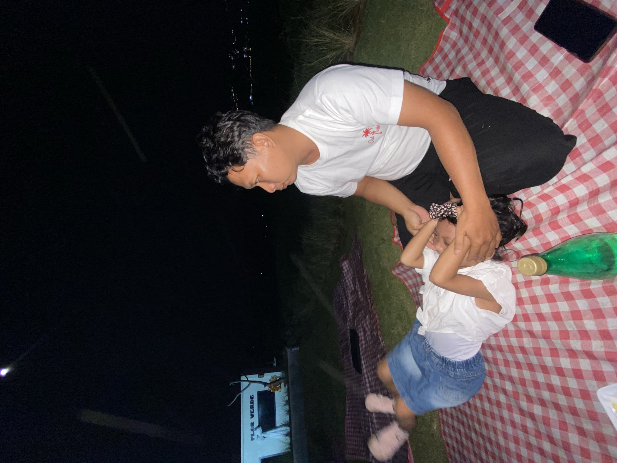 A man and a young child are lying on a red and white checkered picnic blanket spread on grass. The man, wearing a white t-shirt and black pants, is leaning over the child, holding her head. The child, in a white shirt and blue denim skirt, appears to be covering her face. A green bottle and a plate are also on the blanket. In the background, a building with the text "FLOR VERDE" is visible, and there are some lights in the distance suggesting it is nighttime. The scene appears to be outdoors, possibly in a park or recreational area. The lighting suggests it is dark outside, possibly evening or night.