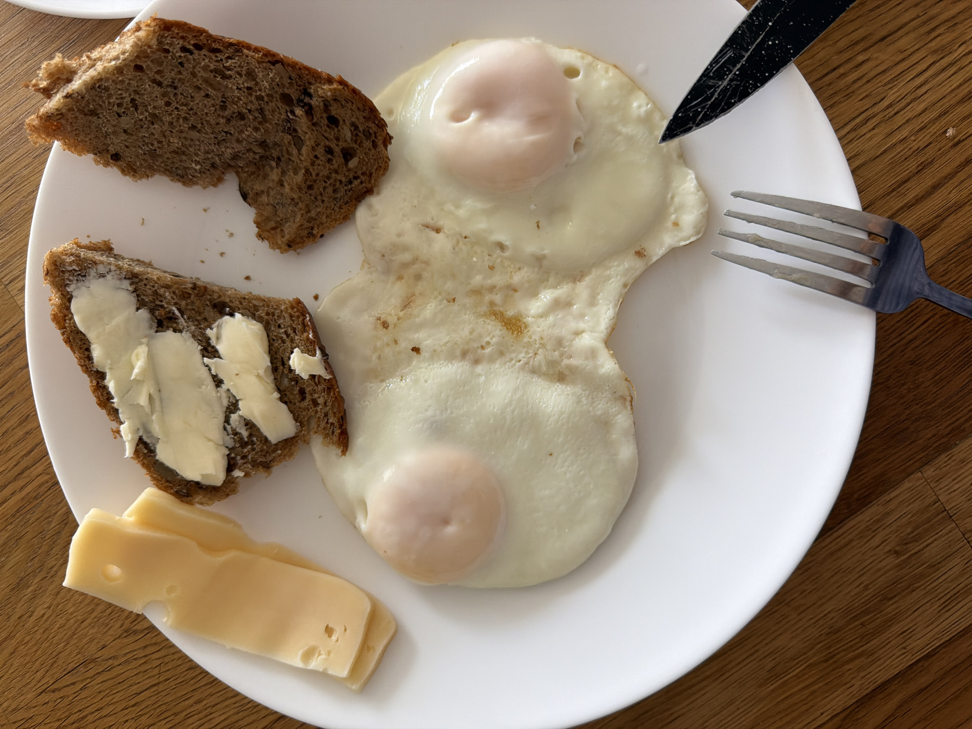 A close-up, top-down shot captures a simple breakfast on a white plate, placed on a wooden table. The meal consists of two fried eggs, two slices of dark rye bread, one topped with butter, and three slices of yellow cheese. A fork and a black knife rest on the plate, ready for use. The scene is lit by overhead light, casting soft shadows on the food and table. The overall impression is one of a humble, home-cooked meal.