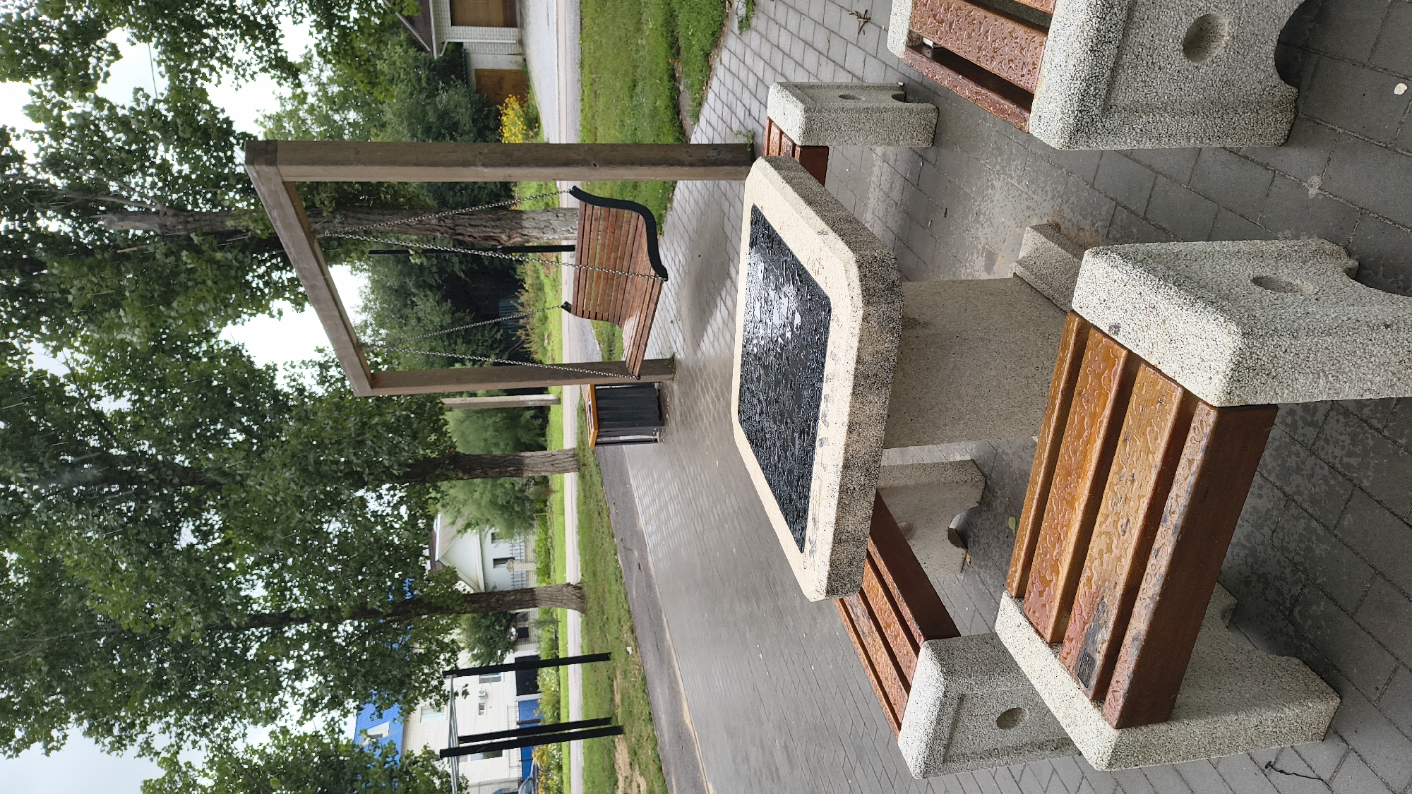 In the city of Mikhaylovka, Russia, a public seating area is depicted under a cloudy sky, with rain visible in the air. The scene features a concrete picnic table with a dark, textured surface that is wet from the rain, surrounded by two concrete benches with wooden slats. To the right, another concrete bench is partially visible. In the background, a wooden swing set with a wooden bench seat hangs from a wooden frame. Several tall trees with green foliage are present, and a pathway paved with grey interlocking bricks leads away from the seating area. Beyond the pathway, a road is visible, with buildings and a small yellow flower bed further in the distance. The overall atmosphere is one of a damp, overcast day in a park or recreational space.