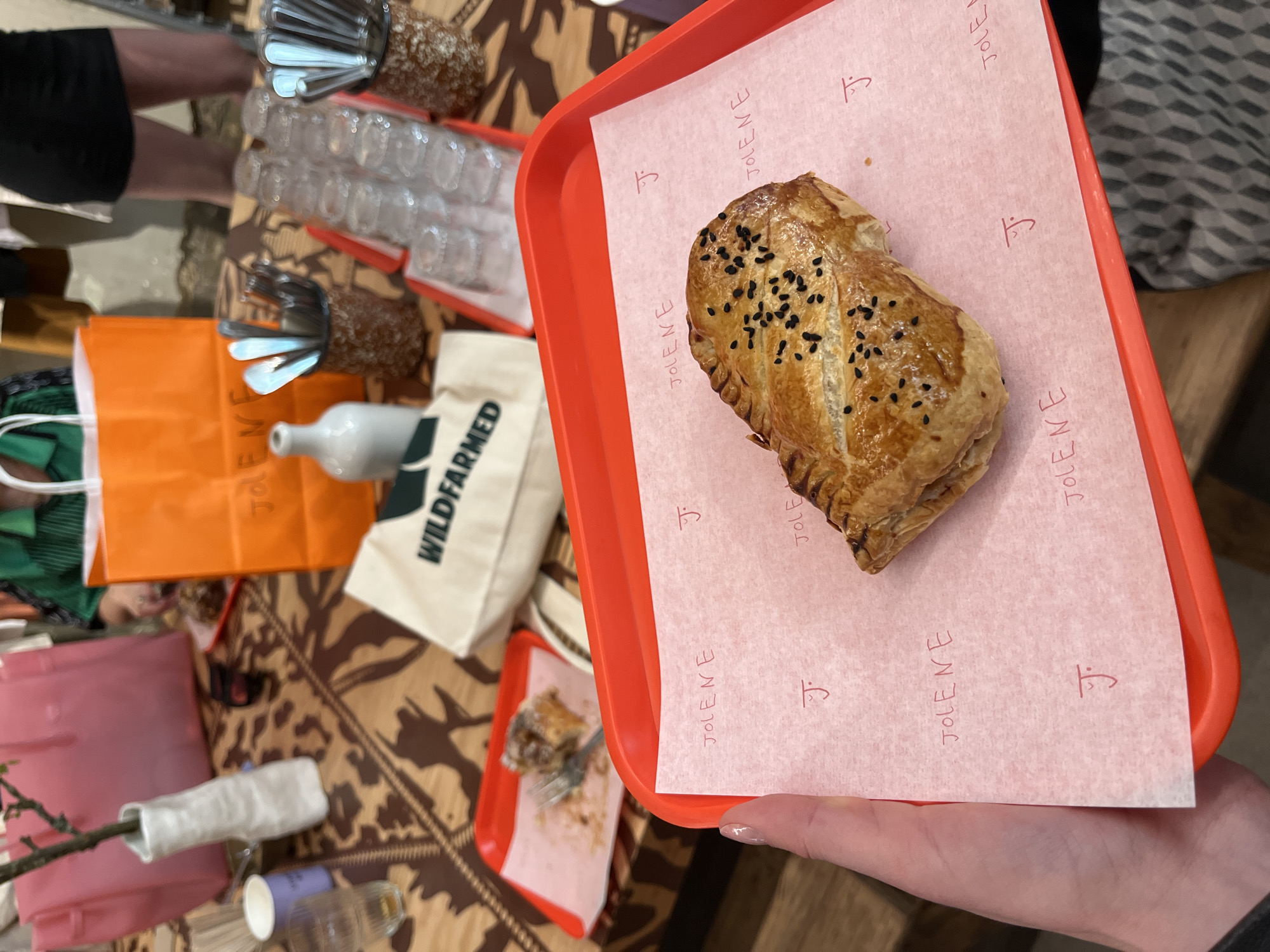 A photograph shows a hand holding an orange tray with a pastry on a pink paper liner that says "JOLENE" in red. The pastry is golden brown and topped with black seeds. In the background, a table with a patterned cloth is visible, along with several clear glasses, a white bottle, shopping bags from "JOLENE" and "WILDFARMED," and a smaller orange tray with remnants of food. The setting appears to be a food establishment in London, United Kingdom, as indicated by the branding on the tray and bags. The shot is taken from above, looking down at the tray.