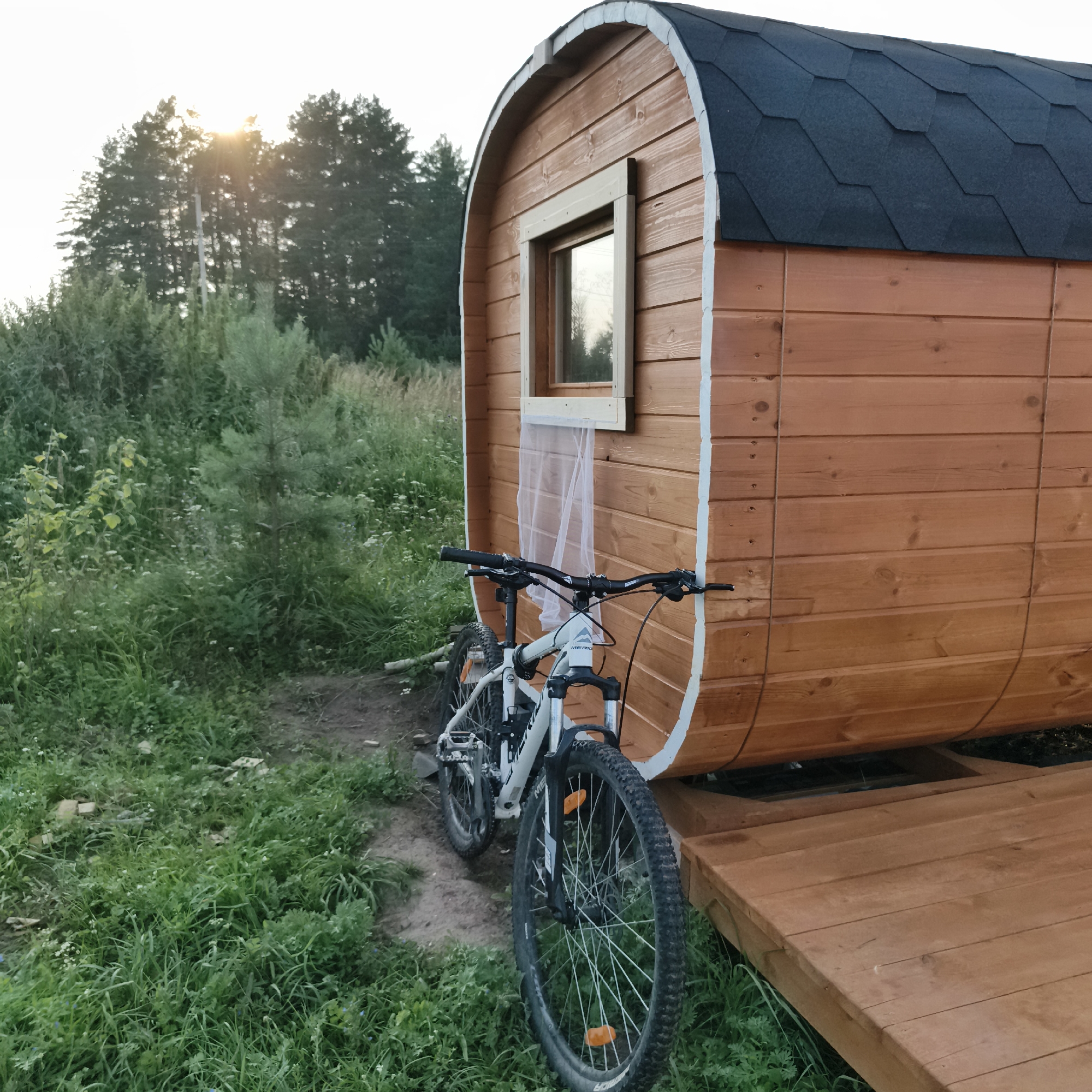 A wooden, barrel-shaped sauna sits on a wooden deck in a grassy area. A white bicycle is parked in front of the sauna. In the background, trees are visible against a bright sky. The sun is setting behind the trees, casting rays of light through the foliage. The scene appears to be located in a natural outdoor setting, possibly a campsite or a rural area. There is no indication of the city of Chaikovsky, Russia in the image.