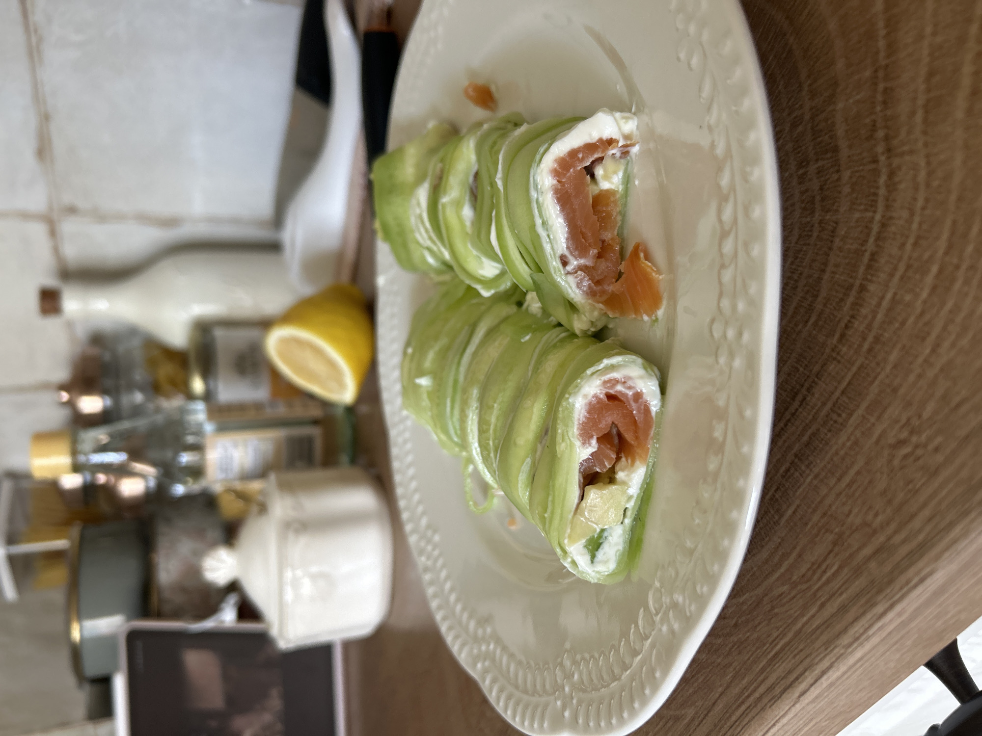 The image shows a close-up of a plate of food on a wooden surface, with a blurred kitchen background. The plate contains two pieces of a dish that appears to be rolled up zucchini or cucumber ribbons filled with cream cheese, pieces of avocado, and smoked salmon. One piece of the rolled dish is cut open, revealing the filling. In the background, there are various kitchen items including bottles of liquid, jars, and a white ceramic sugar bowl. A lemon half is visible on the right side of the frame, and a knife is partially visible behind the plate. The scene appears to be indoors, in a kitchen or dining area. The location of Plovdiv, Bulgaria, is not ascertainable from the provided image.