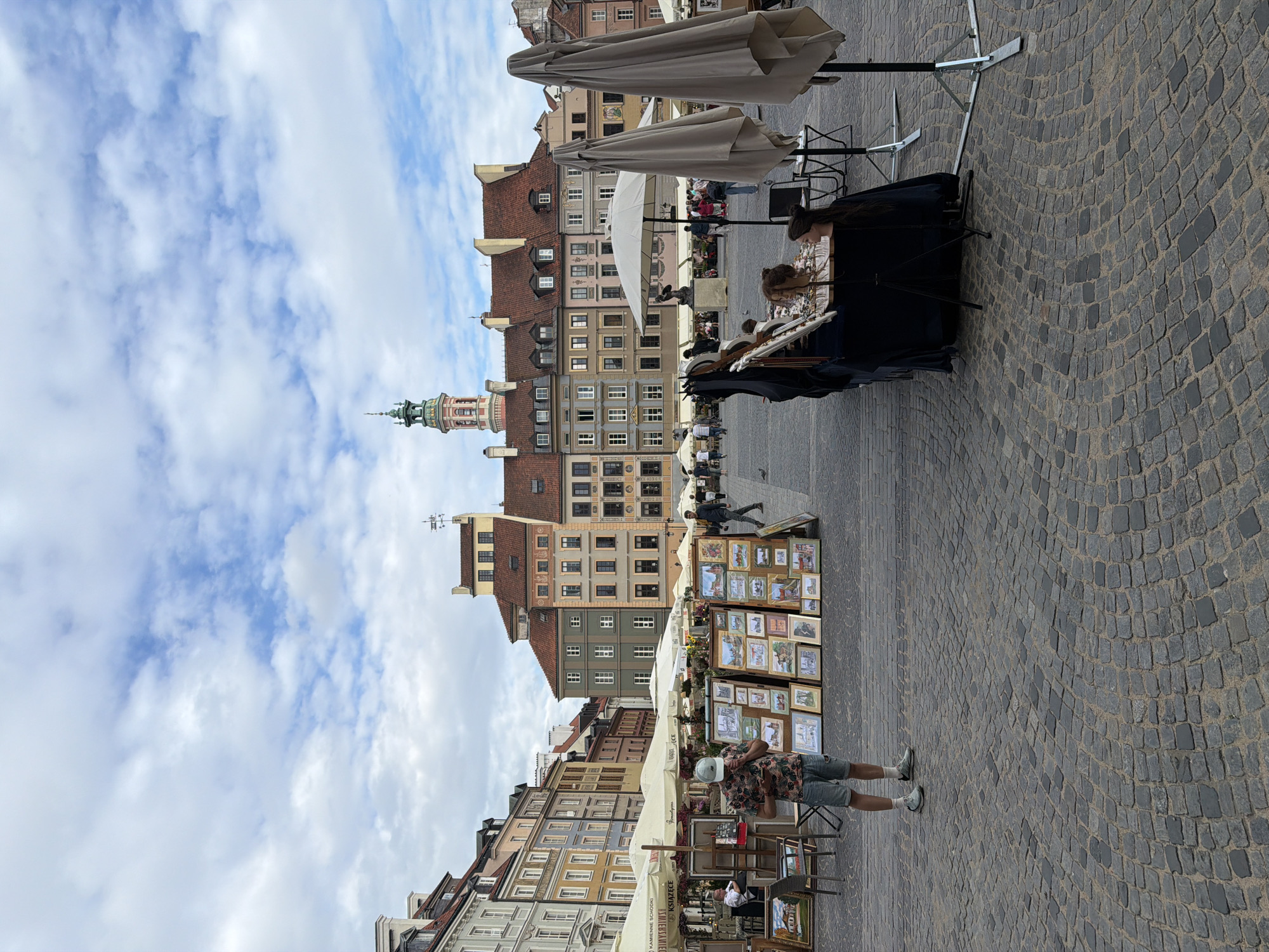 The image depicts a bustling outdoor market scene with people browsing and shopping. The setting appears to be a cobblestone plaza surrounded by colorful, multi-story buildings with traditional architecture. Several stalls are set up, displaying various items, including framed artwork, and some appear to be selling food or drinks under white umbrellas.
A few individuals are visible in the foreground and midground. One man, wearing a patterned short-sleeved shirt and denim shorts, stands near a stall, seemingly looking at something on display. In the background, more people mill about, interacting with vendors and enjoying the atmosphere. Two large, light-colored umbrellas are prominent in the upper part of the image, providing shade over some of the market stalls. The sky is partly cloudy with a mix of blue sky and white clouds, suggesting it's daytime, possibly late morning or afternoon.
The buildings lining the plaza have a European feel, with their pitched roofs, numerous windows, and varied facade colors, ranging from pale yellow and pink to green and brown. A prominent tower with a green spire and clock face rises above the buildings on the left side of the image, possibly a church or clock tower, adding to the historical character of the location. A sign with Polish text, "KAMIENNE SZKOŁY," is visible on a stall. The overall impression is one of a lively and vibrant market in a historic urban setting.