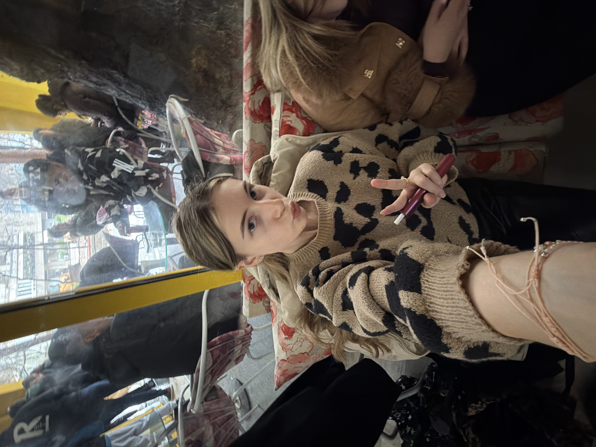 A young woman is posing for a selfie inside a trolleybus in Plovdiv, Bulgaria. She's wearing a leopard-print sweater, black pants, and a bracelet, and is holding a red electronic cigarette. Another person wearing a brown jacket is seated nearby. Through the trolleybus window, a street scene with buildings, trees, and pedestrians is visible. The bus interior includes floral print upholstery.