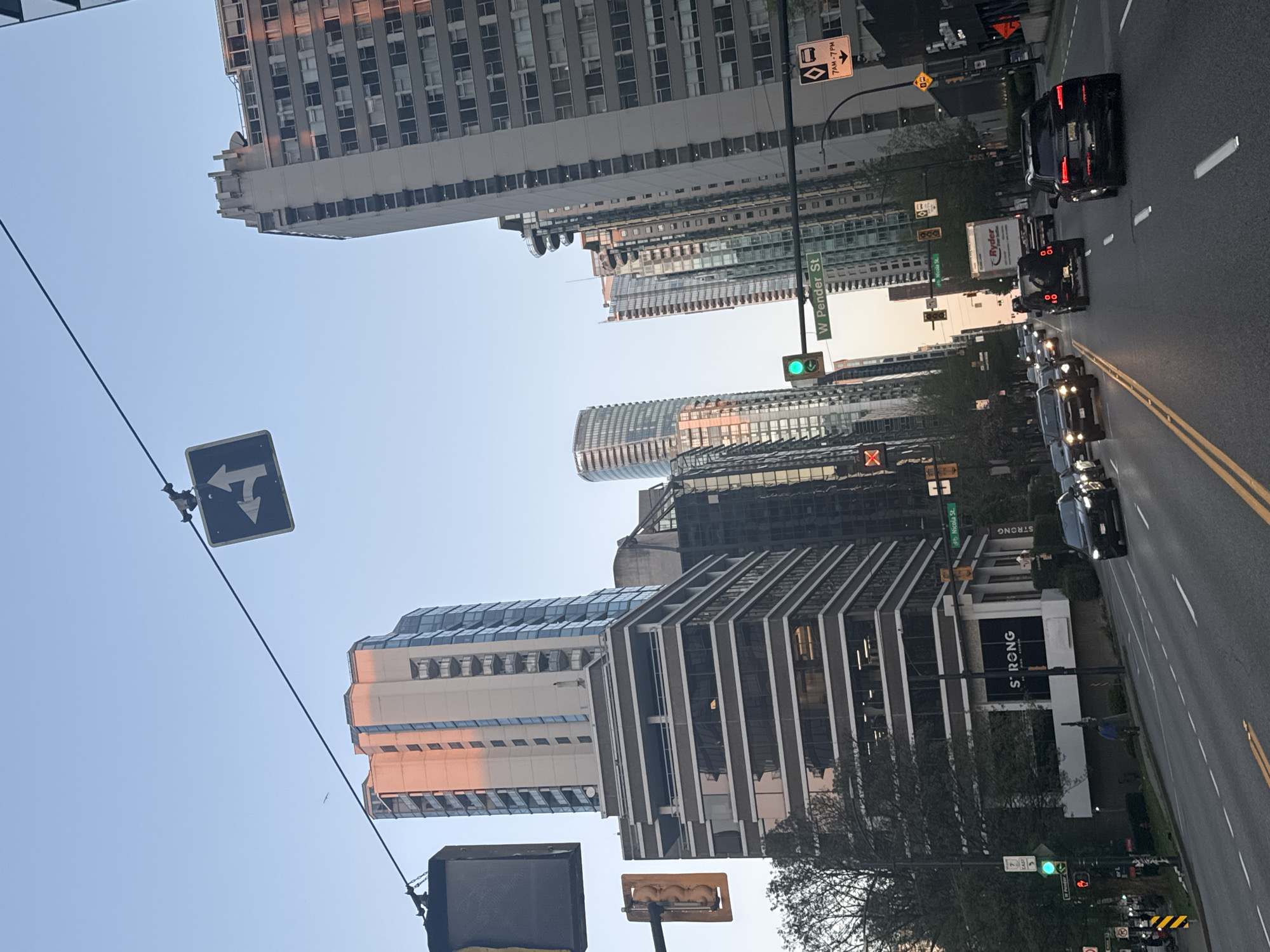 The image captures a vibrant urban street scene in Vancouver, Canada, during what appears to be late afternoon or early evening due to the warm light reflecting on some buildings and the bright, clear sky. The perspective is looking along a street lined with numerous high-rise buildings.
On the right side of the image, the street features multiple vehicles, including dark-colored sedans and SUVs, with a prominent black SUV in the foreground of a traffic lane. Further down the road, a white Ryder truck is visible. The asphalt road is marked with yellow double lines and white lane dividers.
On the left side, an array of towering buildings dominates the skyline. These structures exhibit diverse architectural styles, from modern glass facades reflecting the sky to older concrete designs. Some buildings catch a warm, orange glow from the low-angle sun. One building features a distinctive rounded top, while another showcases strong vertical architectural elements. On the facade of one building, the text "STRONG" is clearly visible, with a partial word "ONGLES" below it. A person wearing a blue top and dark pants is visible on the sidewalk beside this building.
Several street signs provide location and traffic information: a green sign reads "W Pender St," and another smaller green sign indicates "Nicola St." A diamond-shaped sign with a bus icon and "7AM - 7PM" is present, likely indicating a bus lane or restricted times. A black square sign with a white left-turn arrow is also visible. A green traffic light is illuminated, and overhead wires stretch across the scene. The overall impression is that of a bustling city environment with ongoing traffic and pedestrian activity under clear skies.