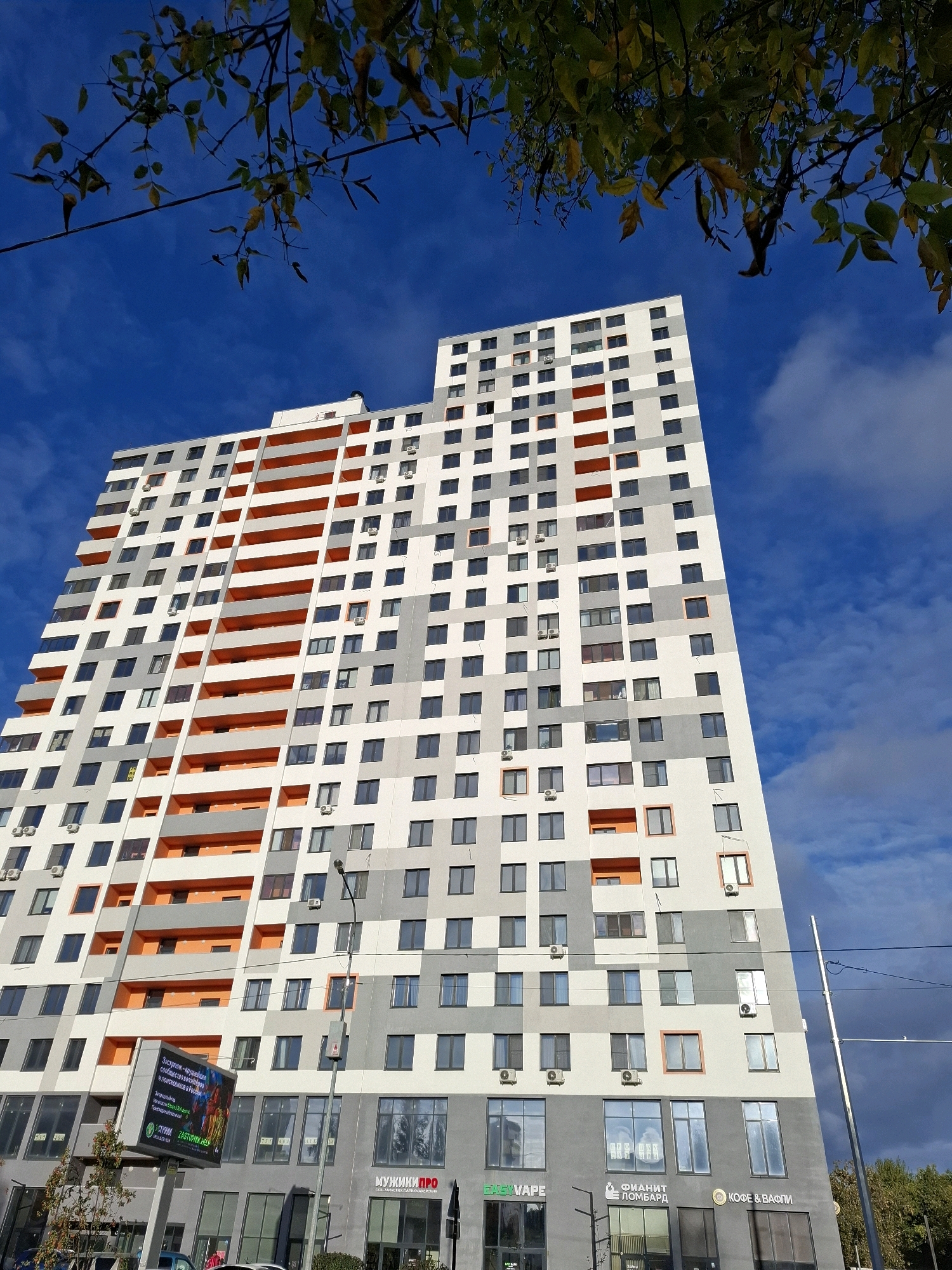 The image shows a modern, multi-story residential building with a distinctive facade featuring alternating white, grey, and orange sections, as well as numerous windows and balconies. The building is situated in an urban environment, as evidenced by the presence of streetlights, power lines, and trees. In the foreground, there is a large digital billboard displaying text and images, and several storefronts with signage are visible at the base of the building. The sky is a vibrant blue with scattered clouds, and the branches of a tree with autumnal foliage frame the top of the image.
The digital billboard displays text in Russian, including "Доступнее - крупнее сообщения золота" and "ZASTUPNIK.HELP". The storefronts also have Russian signage, with names such as "EVAPE", "ФИНАНТ ЛОМБАРД", and "КОФЕ & ВАФЛИ". These elements indicate the location is in Russia.
Based on publicly available information and visual cues, this building is located in Tyumen, Russia. Specifically, this is likely the residential complex "ProNovenko" (ПроНовенко), which is known for its distinctive orange and grey facade and is situated in the city of Tyumen. The digital billboard and the types of businesses at the ground level are consistent with this location.