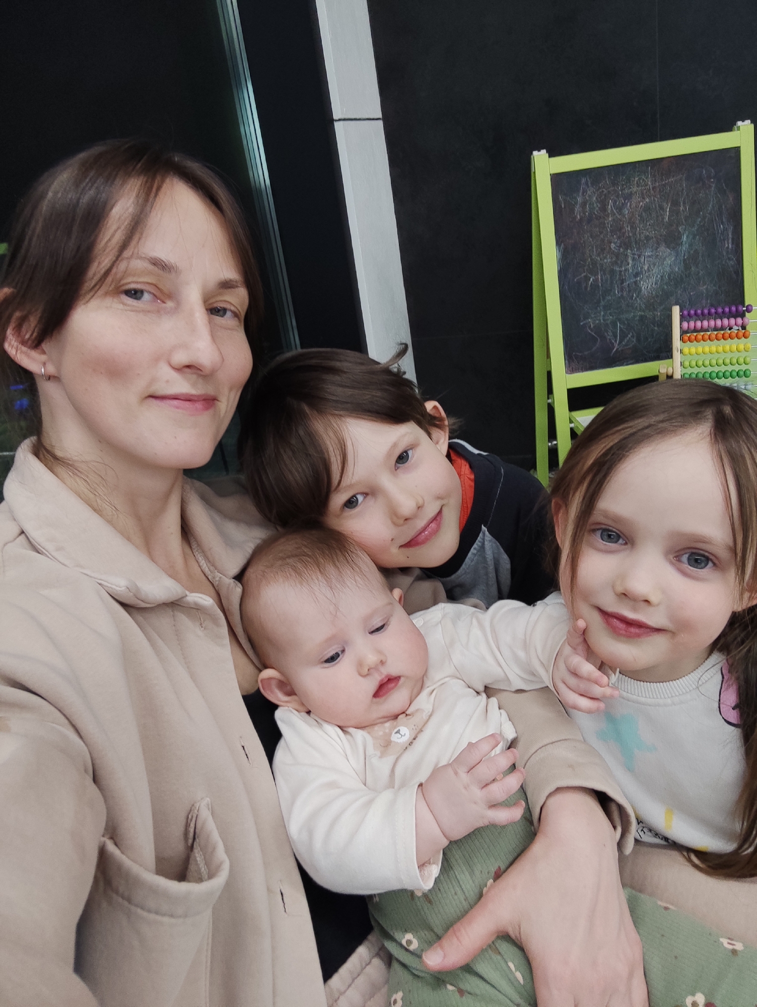 An indoor scene captures four individuals: one adult female, two children (one boy and one girl), and one infant.
An adult female, estimated to be 30-40 years old, with brown hair and light skin, occupies the left side of the frame. She wears a light beige collared jacket or shirt, exhibits a subtle smile, and looks directly towards the camera. A silver hoop earring is visible in her left ear.
A boy, appearing 6-10 years old, with brown hair and light skin, is positioned behind the adult female and slightly to her right. He wears a dark long-sleeved top with a visible orange/red inner collar and smiles towards the camera.
A girl, estimated to be 4-7 years old, with long brown hair and light skin, is at the front-right. She wears a white long-sleeved top with light blue and other indistinct colorful patterns, smiles at the camera, and rests her left hand near her chin.
An infant, under one year old, with light hair and fair skin, is held by the adult female in the center-bottom. The infant is dressed in a light-colored long-sleeved top with a small bow near the collar and green patterned pants. The infant's gaze is directed slightly to the left, away from the camera.
The adult female holds the infant in her arms, her left arm visible supporting the infant's lower body. The adult, boy, and girl all look towards the camera with smiles, suggesting a posed group image.
The background consists of a dark, possibly black or dark grey, wall. To the upper right, a green-framed chalkboard easel is visible, covered in white and colored scribbles. A small, multi-colored abacus with beads in purple, pink, orange, yellow, green, and blue rows is situated on a surface in front of the lower right section of the chalkboard. Vertical white or light-colored structural elements are present on the left side of the background, possibly part of a window or door frame. The setting appears to be an indoor space, likely a home or a child-friendly recreational area.
The specific location within the city of Izhevsk, Russia, cannot be determined from the visual information provided in the image.