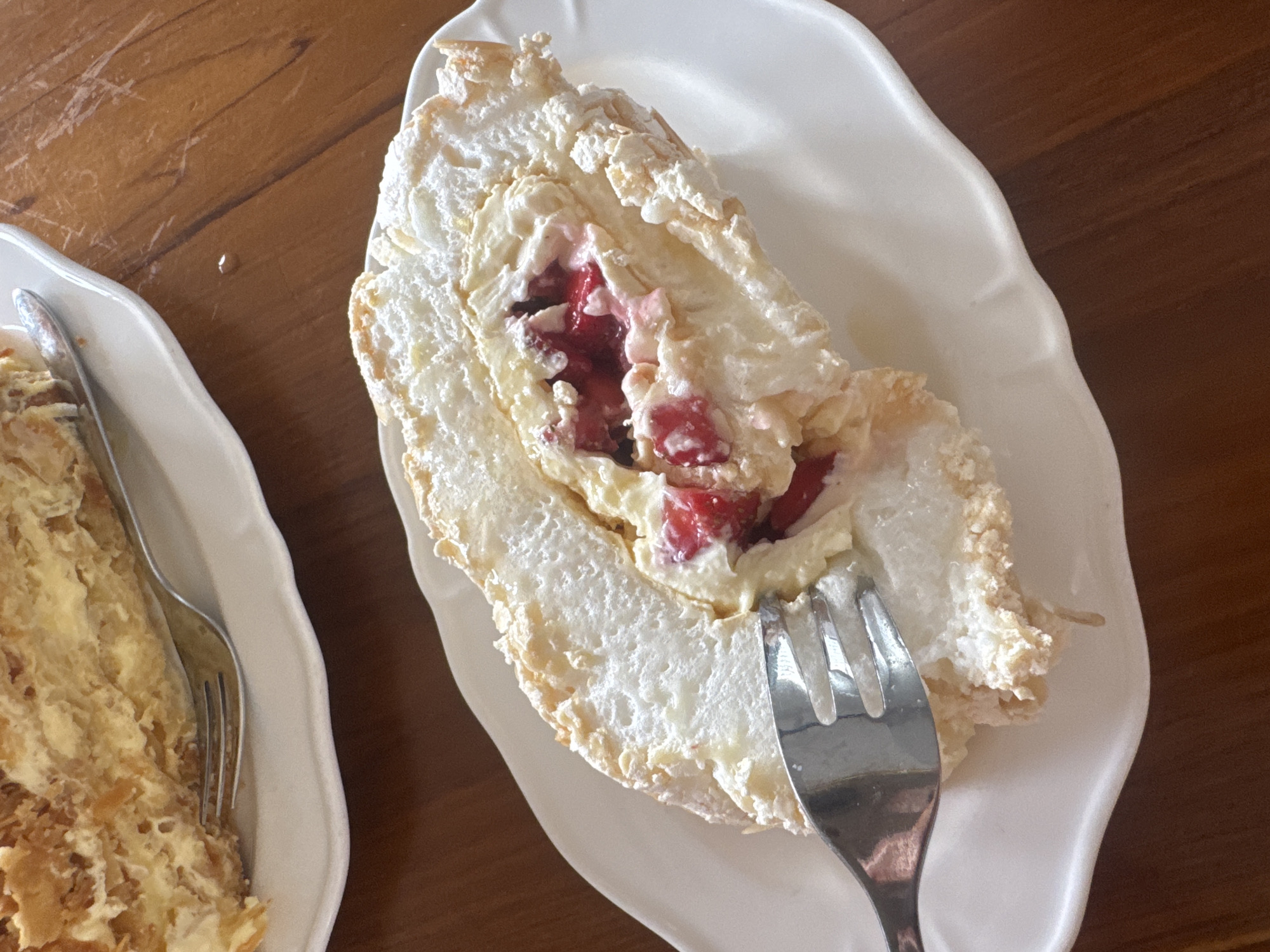 A fork is taking a bite out of a slice of roulade cake with meringue and berries, on a white plate on a wooden table. The roulade cake is light and airy, with a fluffy meringue exterior and a creamy filling studded with red berries. The fork is positioned to grab a piece of the cake. In the background, another slice of cake, possibly a mille-feuille, is visible on another white plate. The setting appears to be a cafe or restaurant in Kuta Selatan, Indonesia, suggested by the warm lighting and the wooden table. The overall scene depicts a sweet treat being enjoyed.
