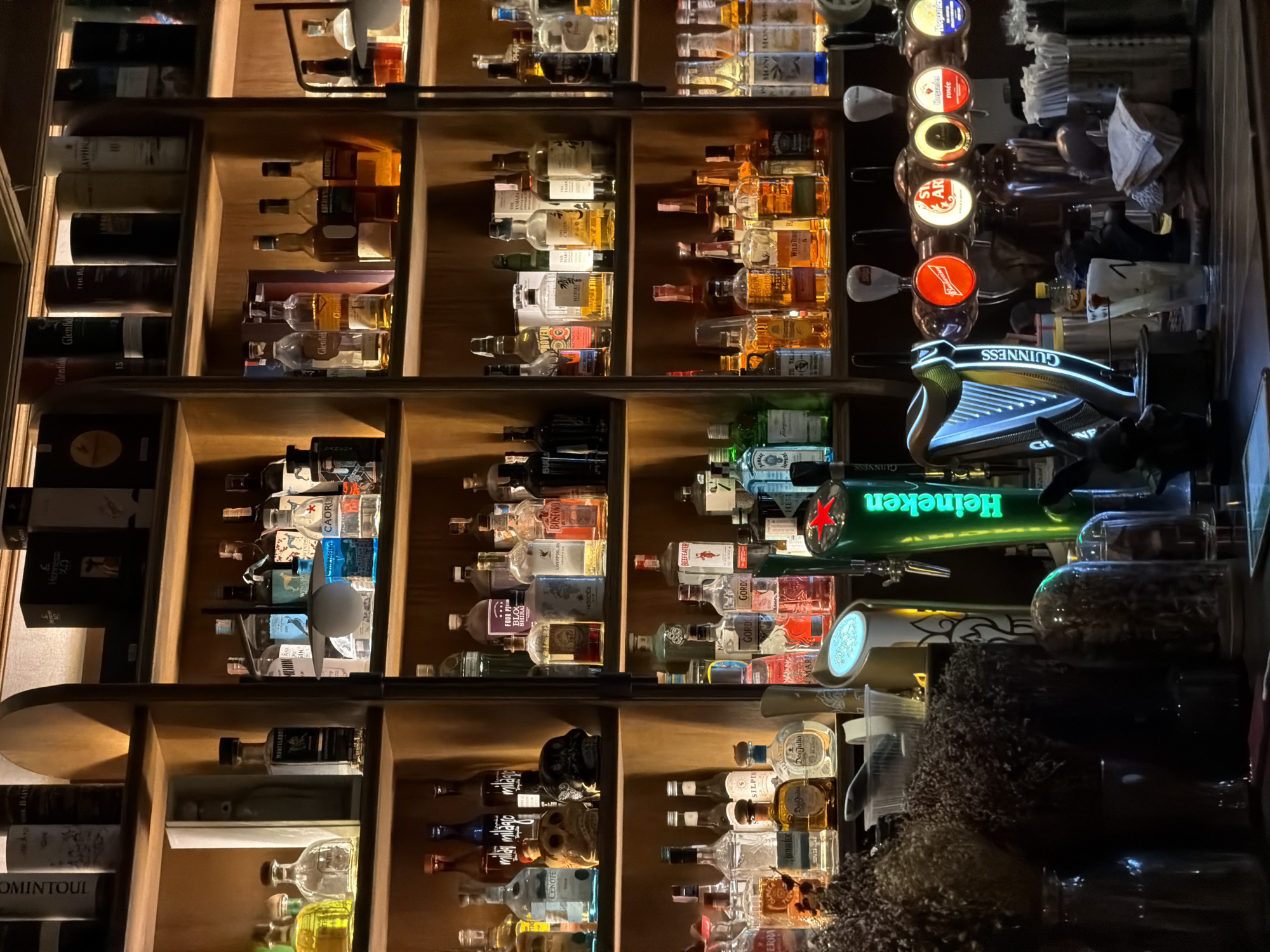 The image depicts a dimly lit bar with shelves stocked with an assortment of alcoholic beverages. The shelves are made of light-colored wood and are organized into sections. On the shelves, numerous bottles of liquor, wine, and other spirits are visible, with labels indicating brands such as Glenfiddich, Caorunn, Hennessy, and many others. Some bottles are clear, showcasing the color of the liquid inside, while others are opaque.
In the foreground, there is a bar counter with several beer taps. A prominent Guinness tap with a harp design is visible on the left, glowing with blue neon lights. Other taps, some illuminated with red and yellow lights, display logos for brands like Budweiser and Stella Artois. Various glasses, some containing drinks, are arranged on the counter. Decorative items, including dried flowers and small figurines, are also present on the counter and shelves. The overall ambiance suggests a typical bar setting, likely in a city given the variety and quantity of alcoholic beverages displayed.
Without further information, it is not possible to definitively pinpoint the location as specifically "บ้านบางรักษ์, Thailand." The image itself does not contain any explicit indicators of this particular location.