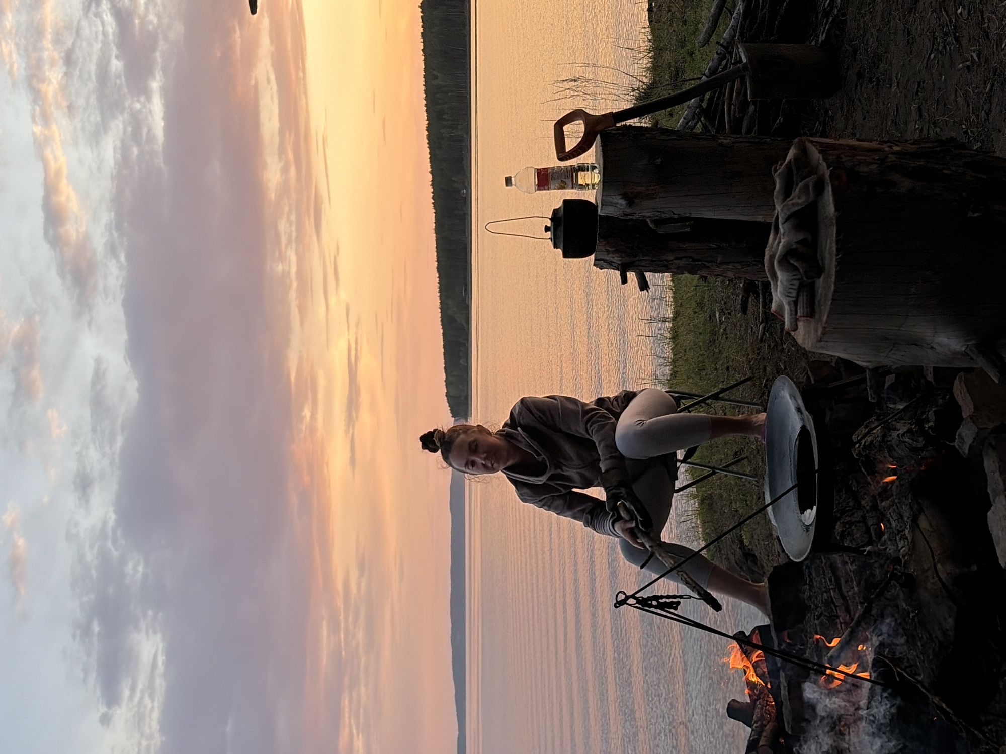 A woman is sitting near a campfire with a large metal lid in her hands near a lake at Zabegaevo, Russia. The sunset colors are reflected on the lake. A kettle and water bottle sit on a nearby log, along with a shovel.