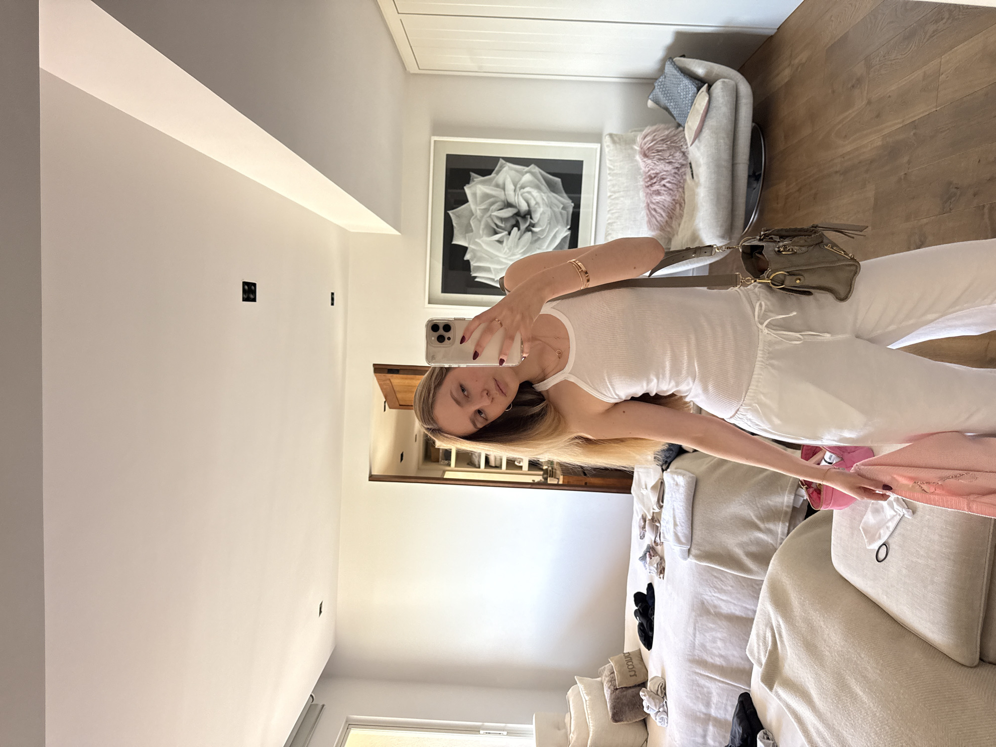 The image depicts a woman taking a mirror selfie inside a well-lit, modern residential bedroom located in Mexico City, Mexico. The woman, with long light-brown hair, is positioned centrally, holding a silver-colored iPhone (likely a 14 Pro Max model due to its camera array) in her left hand, which is visible in the mirror's reflection. She wears a white ribbed tank top and white drawstring pants. Her left hand displays rings on the ring and index fingers, and her right wrist features multiple gold bracelets. A grey crossbody bag with gold hardware is slung over her left shoulder. In her right hand, she holds a light pink garment, possibly a sweater.
The bedroom features white walls, a white ceiling with recessed lighting, and light brown hardwood flooring. To the left, a large bed with light-colored linen bedding is visible, adorned with multiple pillows, including one dark brown pillow with reversed text. Various items of clothing, including folded black garments, socks, and a folded white towel, are arranged on the bed. In the foreground, a light beige upholstered ottoman or bench holds a black hair tie, a small white drawstring bag, and a bright pink zippered pouch containing personal items.
In the background, a light grey swivel armchair with textured fabric is situated to the right of the bed, featuring a pink faux fur pillow and a blue patterned cushion. Above the armchair, a large black and white framed photograph of a multi-petaled flower (resembling a rose) hangs on the wall. To the far left, a vertical window with a white frame and a partially visible beige roller blind allows natural light into the room. A dark wooden door, slightly ajar, leads to an adjacent room, revealing shelves and storage within. White built-in wardrobe doors are visible on the far right side of the room.
