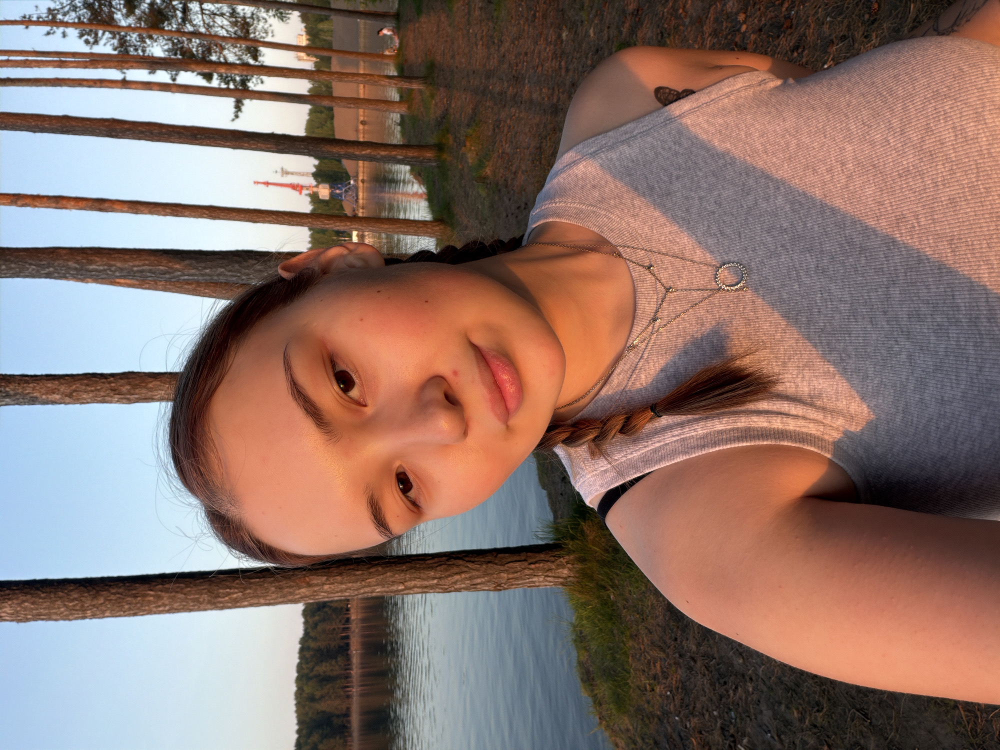 A young woman poses for a selfie by a river or lake. She has brown hair styled in braids, a fair complexion, and wears a grey tank top with a delicate silver necklace. A small tattoo is visible on her left shoulder. The background shows a row of tall, thin pine trees lining the riverbank, with a body of water and distant buildings visible through the trees. The scene is bathed in warm, golden light, suggesting either sunrise or sunset.
Based on the presence of a distinctive red and white structure visible in the distance, this location is likely in the city of Chaikovsky, Russia. The structure appears to be a part of an industrial or recreational facility along the Kama River.