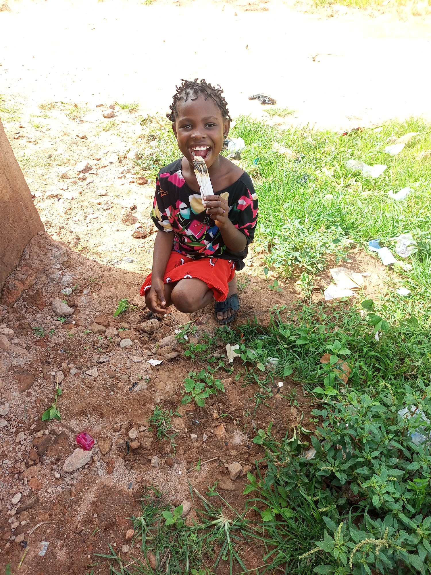 A young Black girl with braided hair is captured mid-action, squatting on dry, reddish-brown earth next to a patch of green grass. She is holding a white object, possibly food, to her mouth and is looking directly at the camera with a wide, joyful smile, revealing her teeth. Her attire consists of a colorful, patterned short-sleeved shirt and a vibrant red skirt. She wears dark sandals. The background shows more dry ground with scattered rocks and some green vegetation, including a prominent patch of grass to her right, which contains some white plastic litter. To the left of the frame, a portion of a rough, reddish-brown wall or structure is visible. The scene appears to be outdoors, possibly in a yard or an undeveloped area within Tempure, Nigeria.