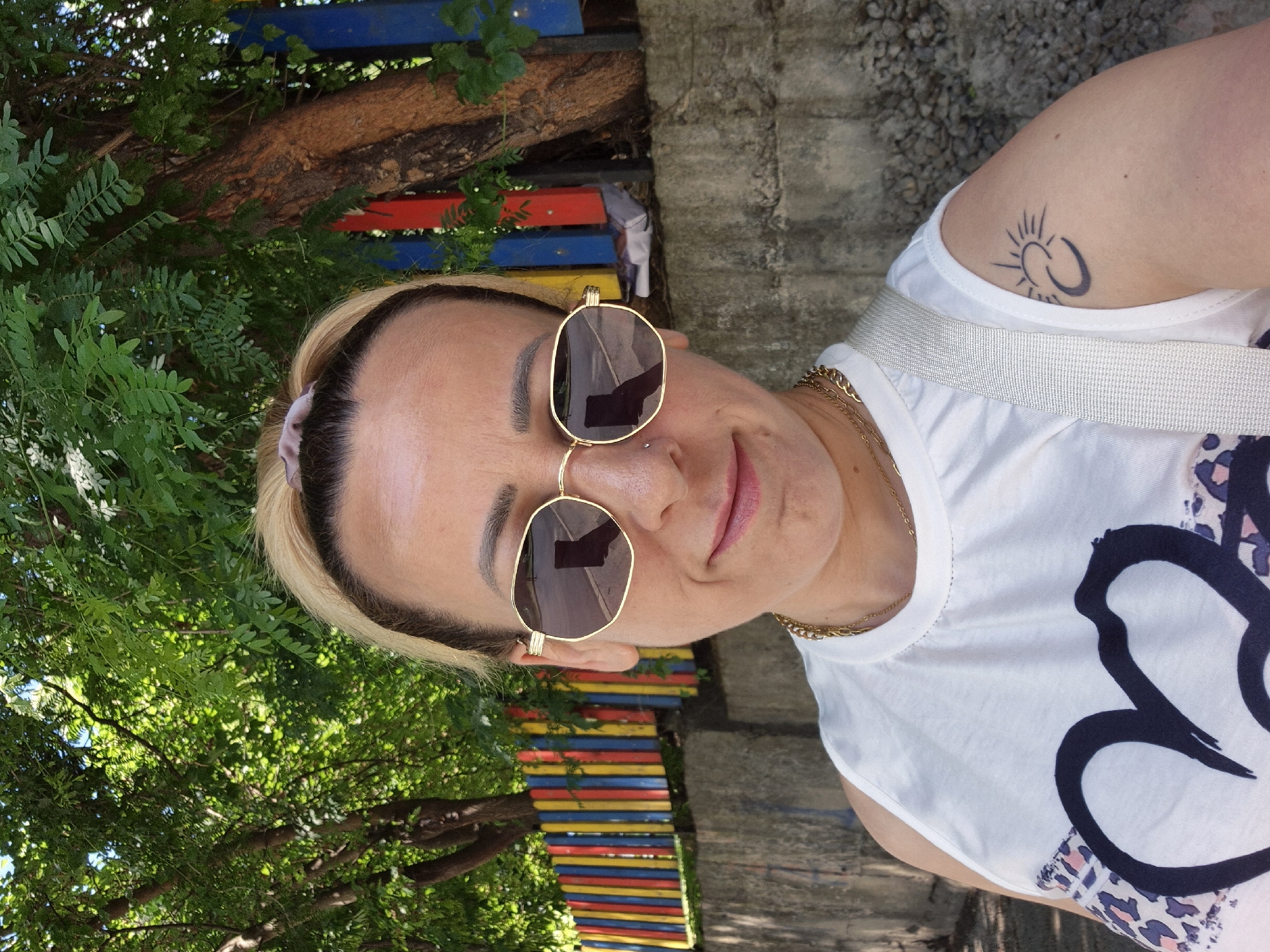 A woman with blonde hair pulled back with a scrunchie, wearing gold-rimmed sunglasses, a white sleeveless shirt with a large dark heart graphic and leopard print accents, and a gold chain necklace, poses for a selfie outdoors. She has a tattoo of a sun and crescent moon on her right arm. In the background, a colorful wooden fence with vertical red, yellow, blue, and green posts is visible, along with trees and a concrete wall. The image appears to be taken in a city setting, possibly a park or schoolyard, on a sunny day.